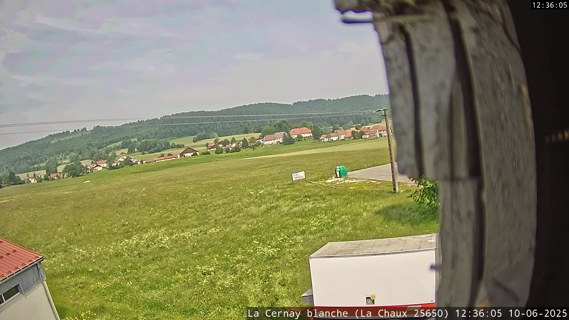 A mostly sunny day shows a wide green field with a small village and forest in the background, viewed from a slightly elevated position.