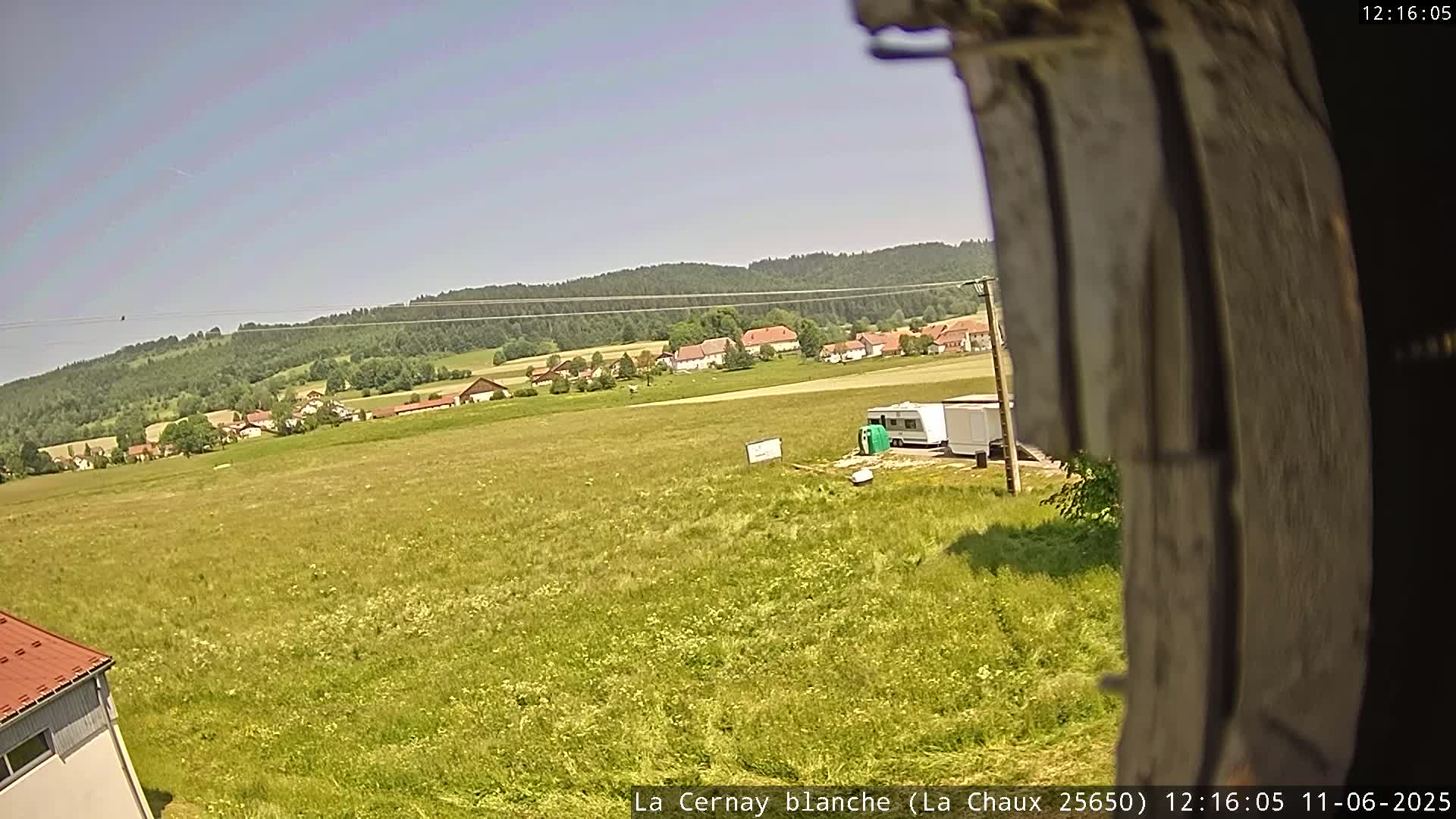A wide, sunny view of a grassy field with a small village and forested hills in the background, showing a camper parked near a building in the foreground.