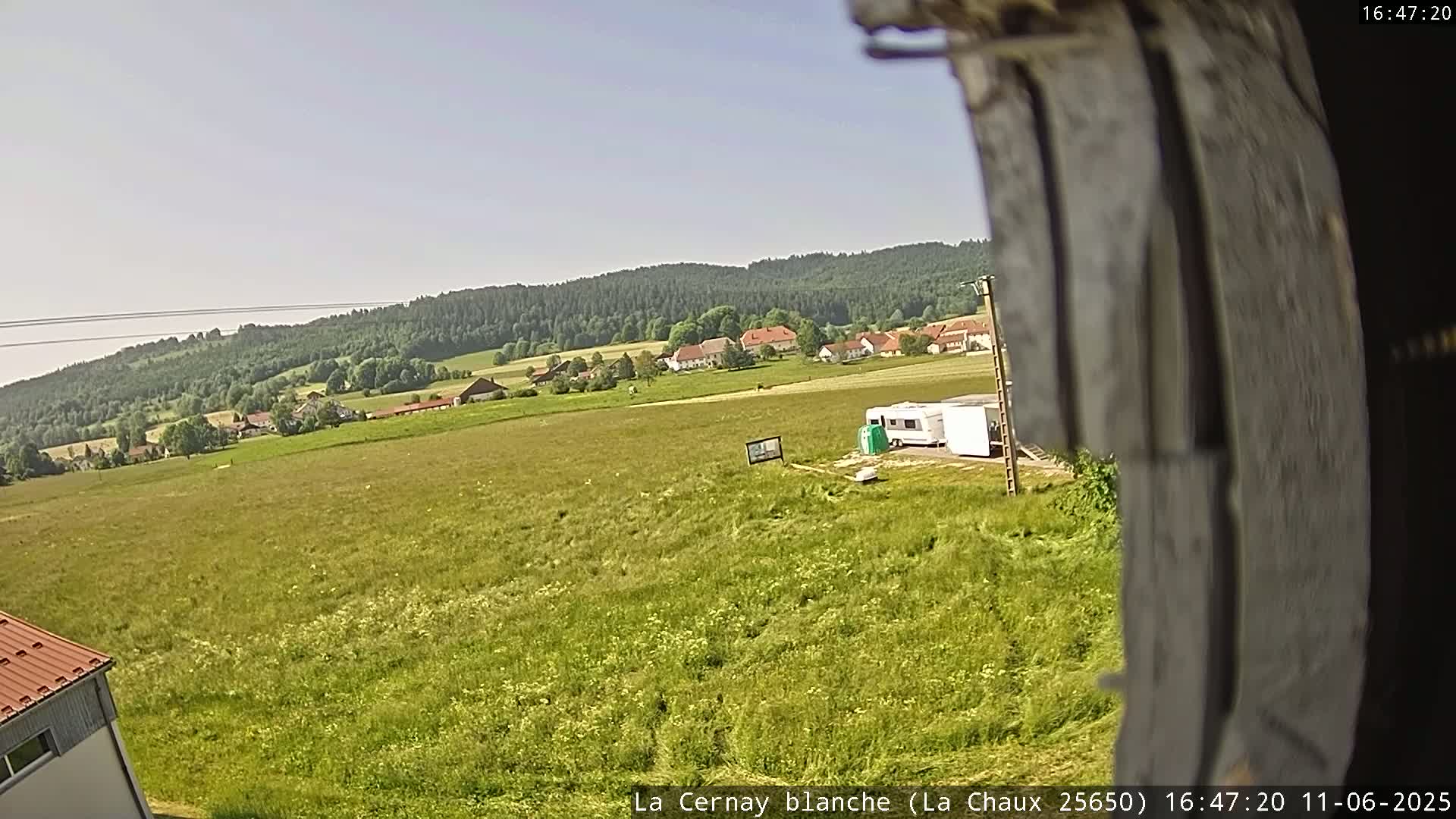 A large grassy field stretches out before a small village nestled in a valley surrounded by a forest under a clear sky.