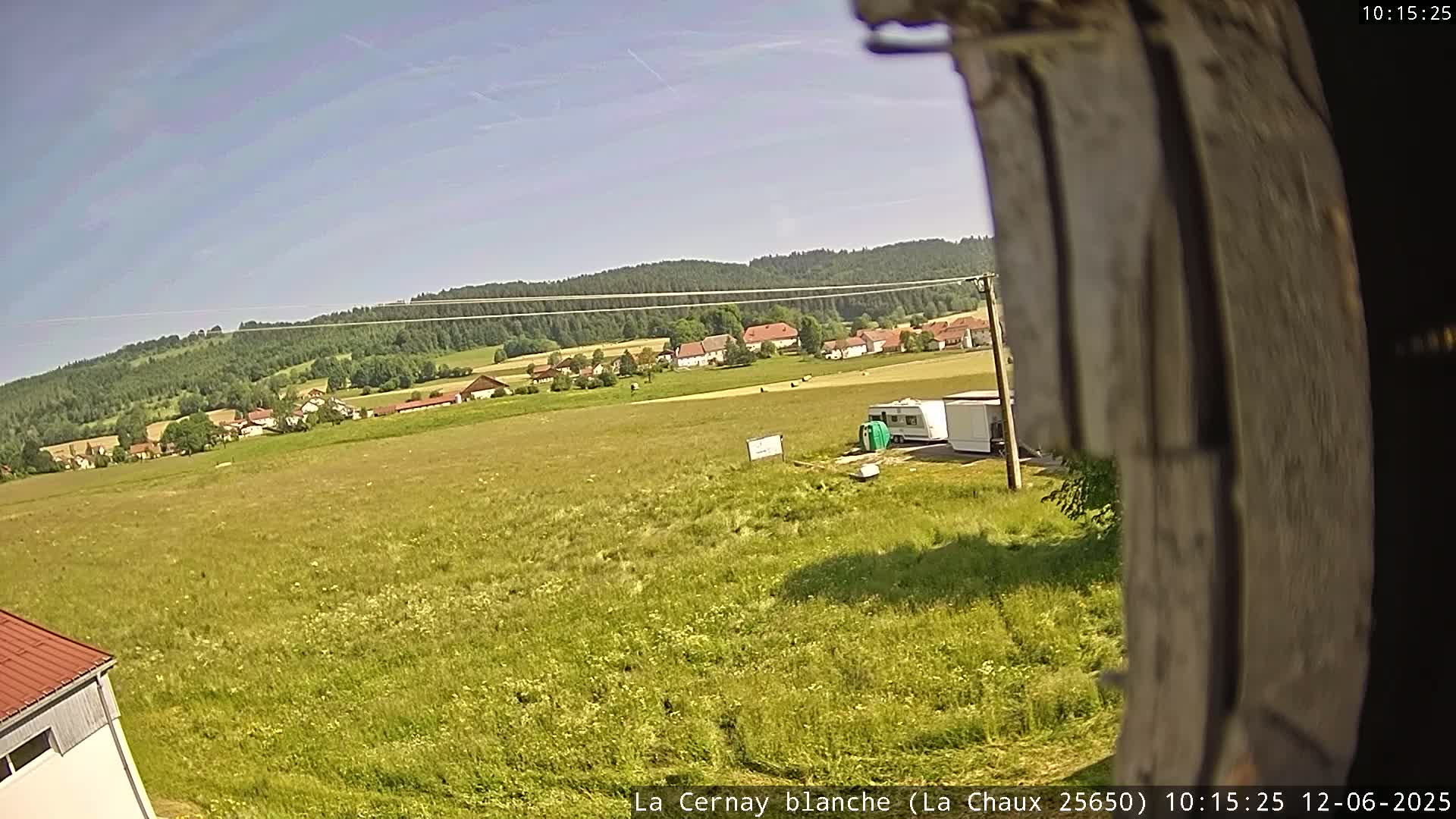 A wide, sunny field stretches out before a small village nestled against a verdant hillside under a clear blue sky.