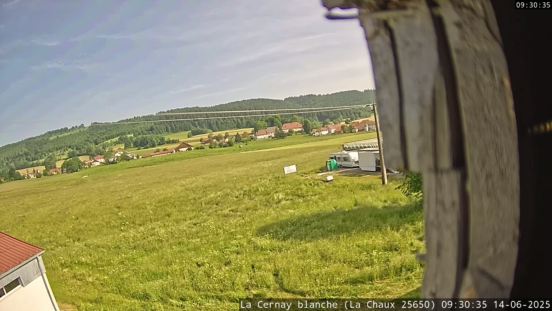 A wide, sunny shot shows a field of green grass with a small village and forested hills in the distance, and a few vehicles parked near the bottom of the frame.