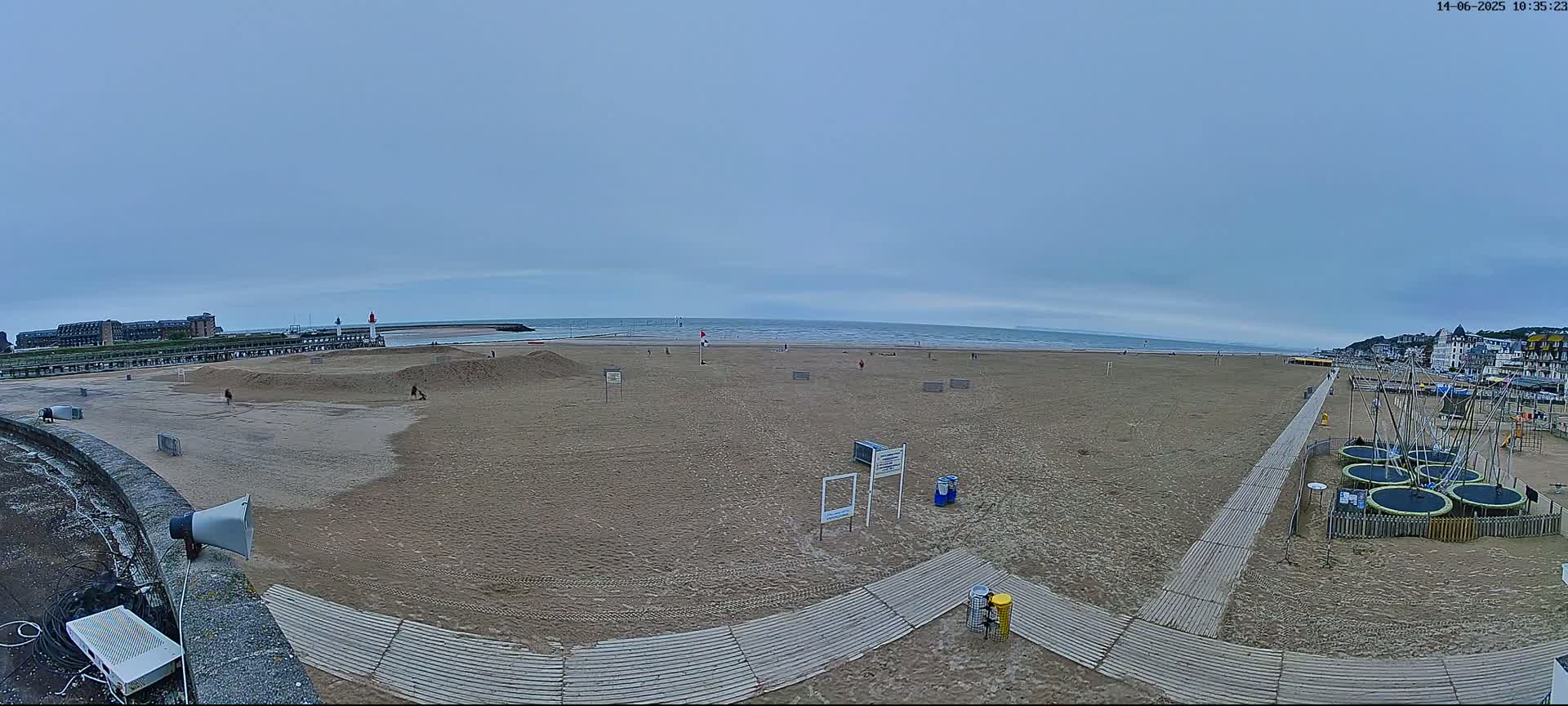 A wide shot of a mostly empty sandy beach on an overcast day, with a calm sea in the background, several small structures and some trampolines on the right, and a few people scattered across the sand.