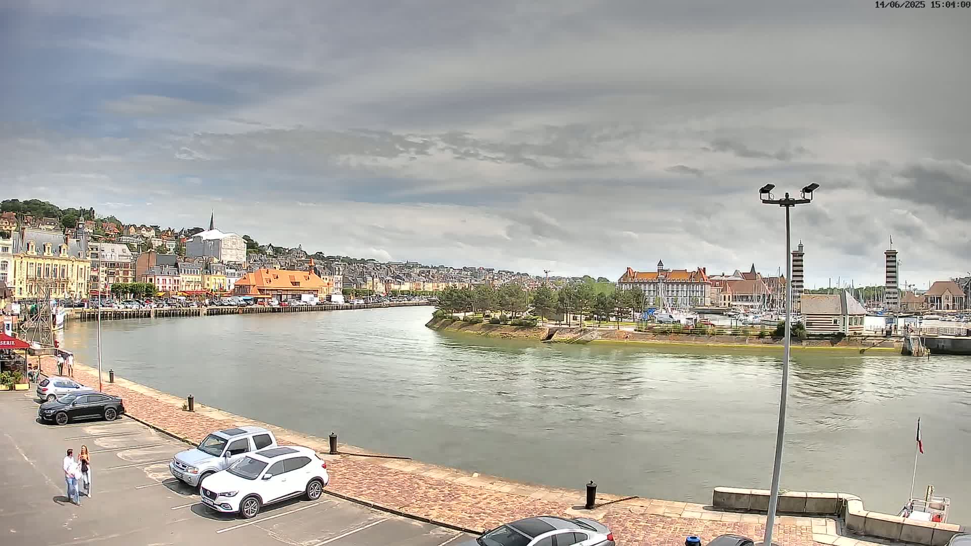 A river flows through a town with colorful buildings under a cloudy sky, cars parked in a lot alongside the river.