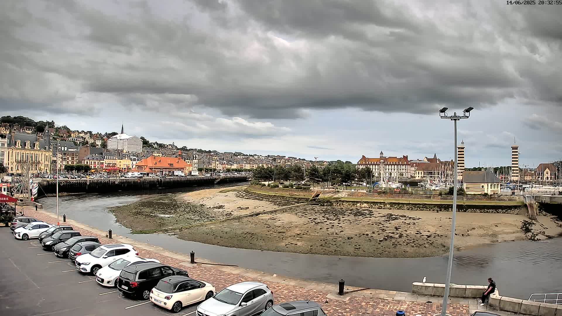 A river flows through a town with colorful buildings under a cloudy sky, cars parked in a lot alongside the river.