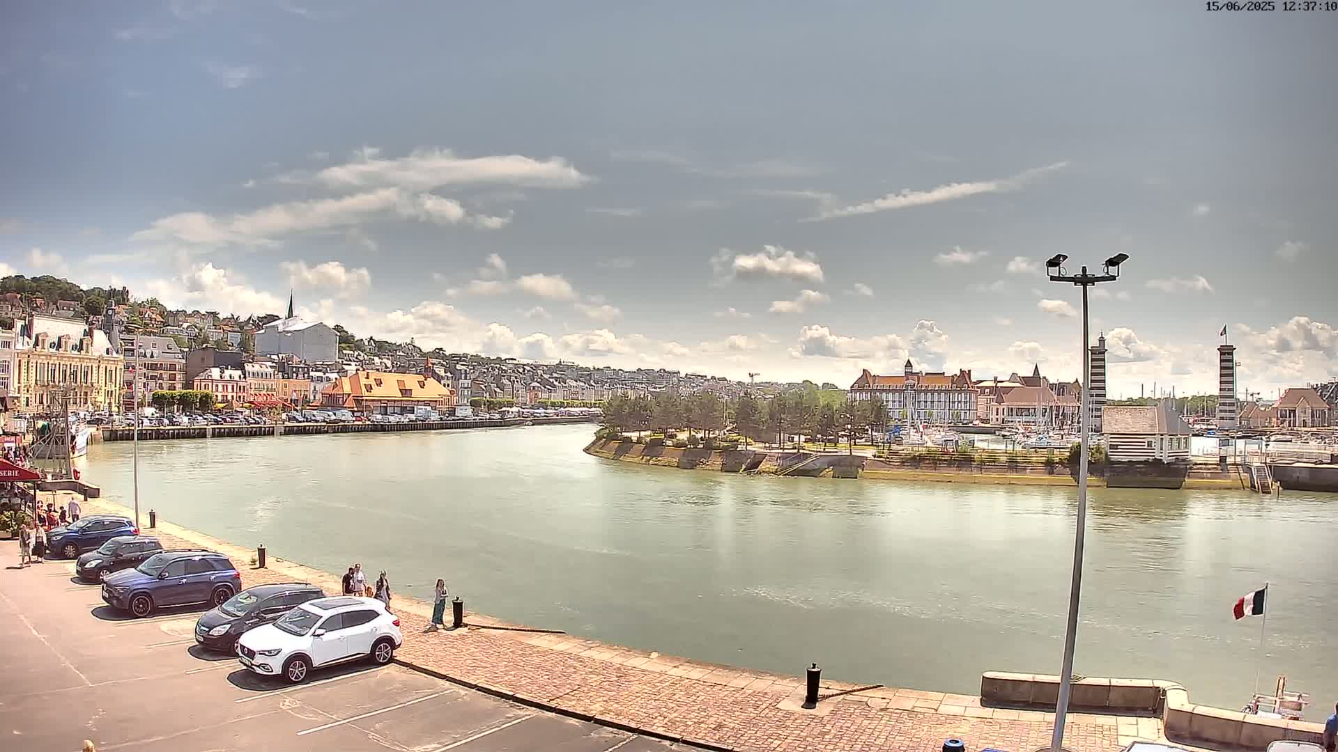 A partly cloudy summer day shows a river curving past a town with buildings, parked cars, and a marina.