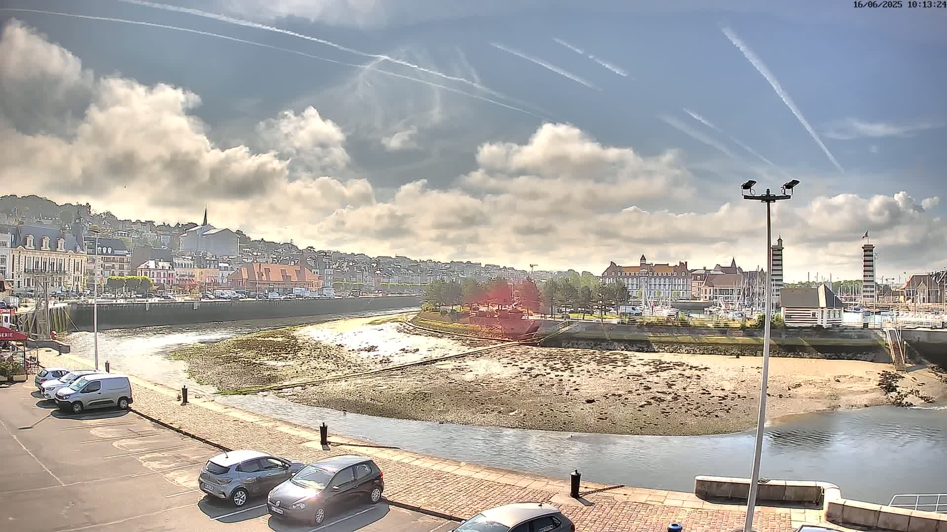 A partly cloudy sky overlooks a harbor town with a low-tide river curving around a sandy area, several cars parked along the waterfront, and a line of buildings in the background.