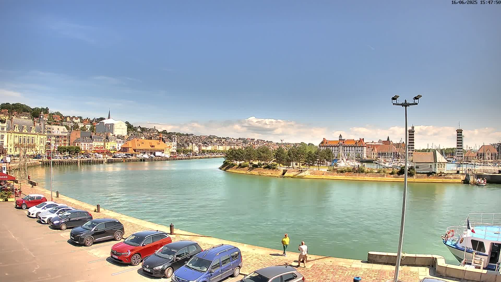 A sunny day reveals a harbor town with colorful buildings lining a calm waterway, where several cars are parked and a few people stand by the water's edge.