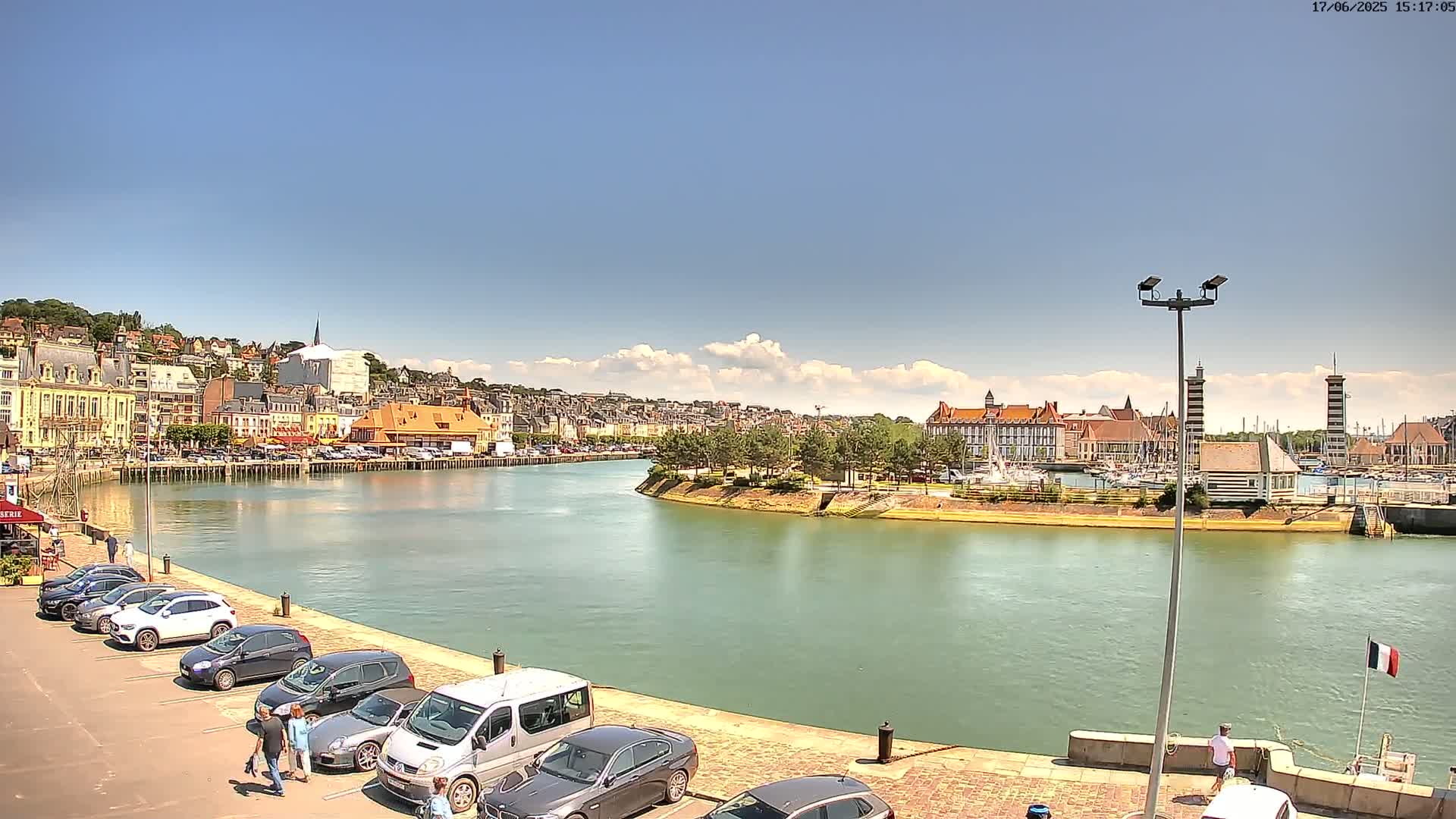 A sunny day reveals a harbor town with a calm waterway curving around a green landmass, lined with buildings and cars parked along the quay.