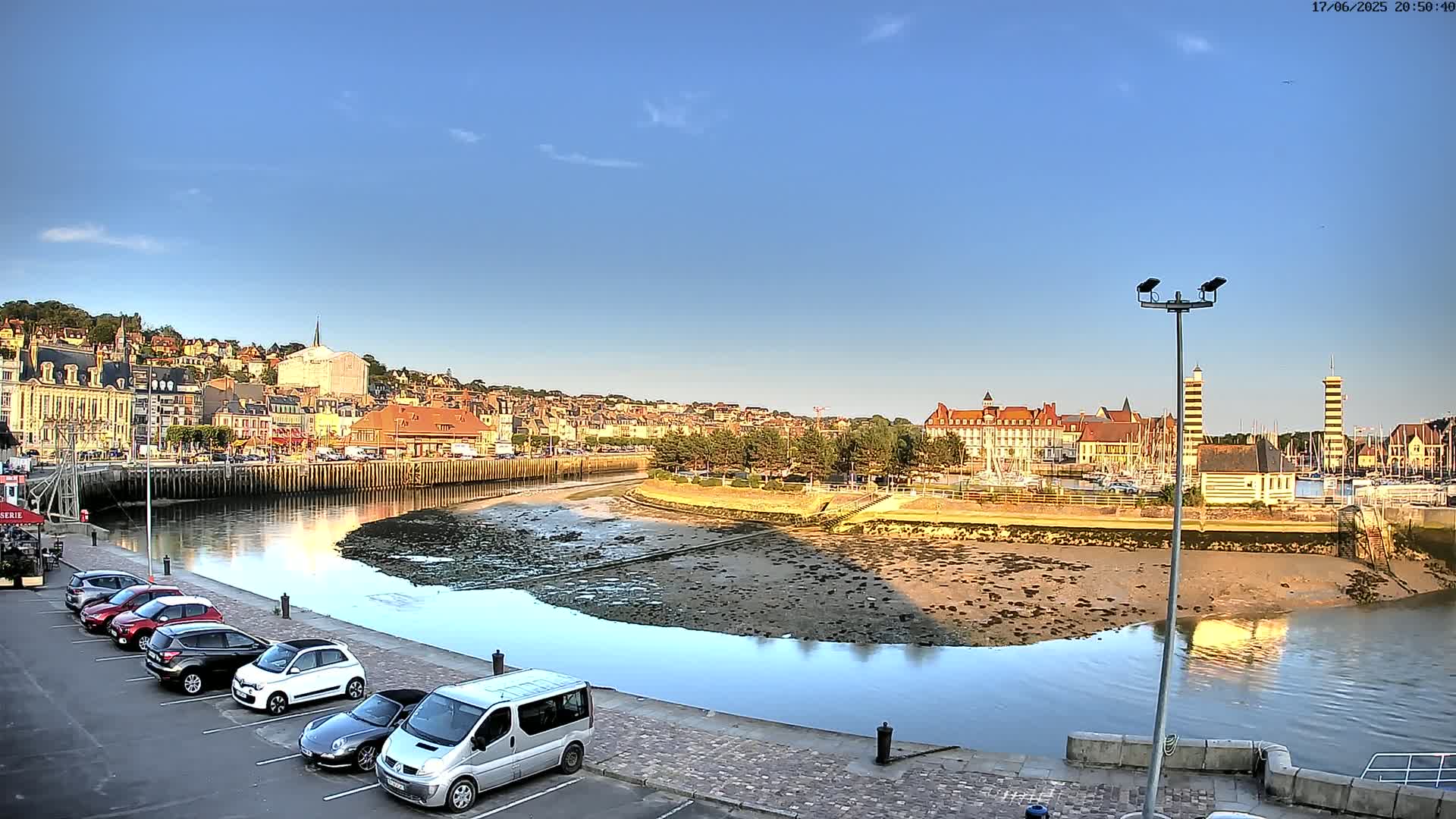 A harbor scene on a sunny day shows a calm waterway with exposed mudflats, a parking lot with several cars, and a town built on a hillside in the background.