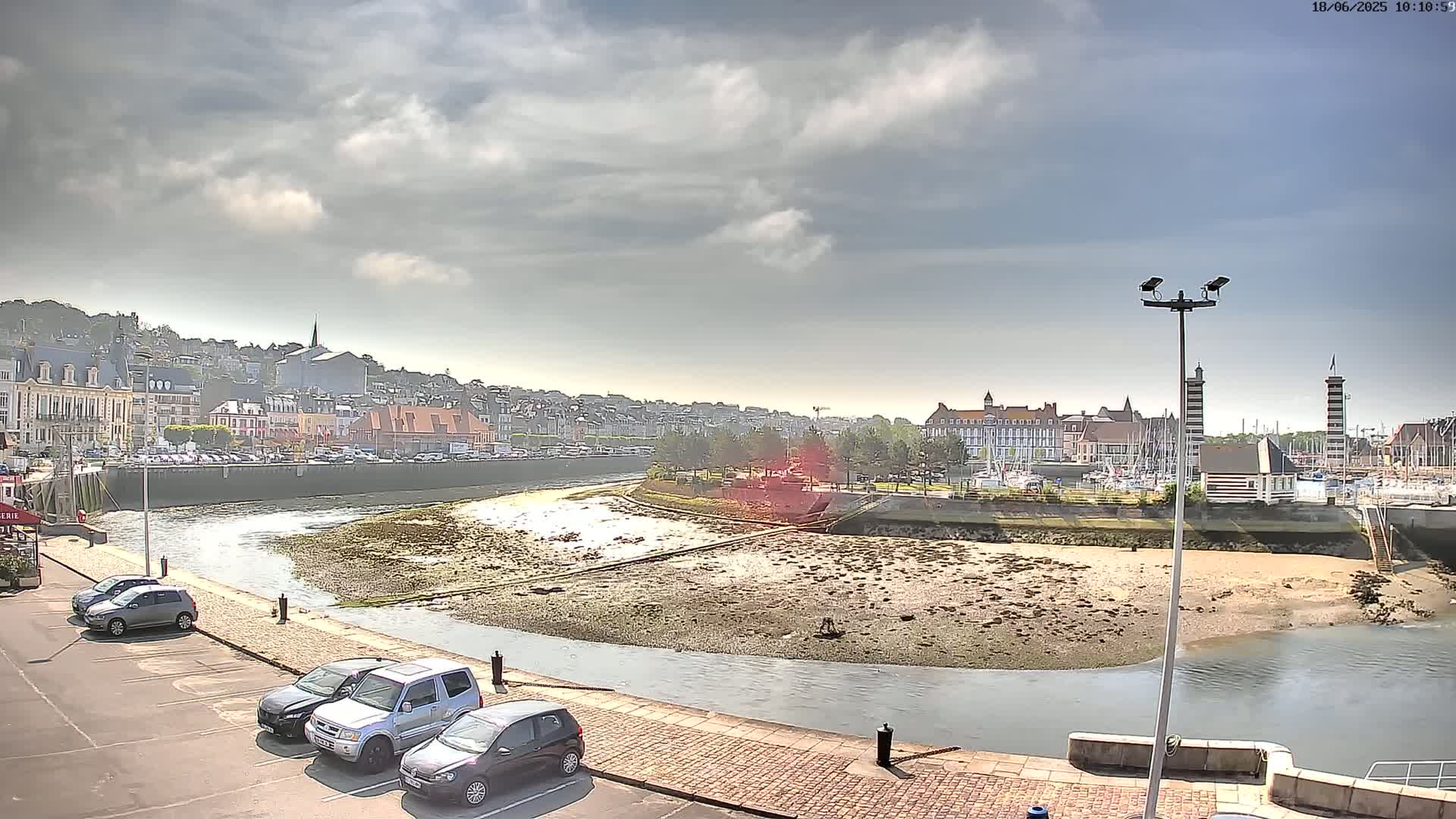 A mostly sunny day reveals a harbor scene with a partly exposed riverbed, several parked cars, and a town built on a hillside in the background.