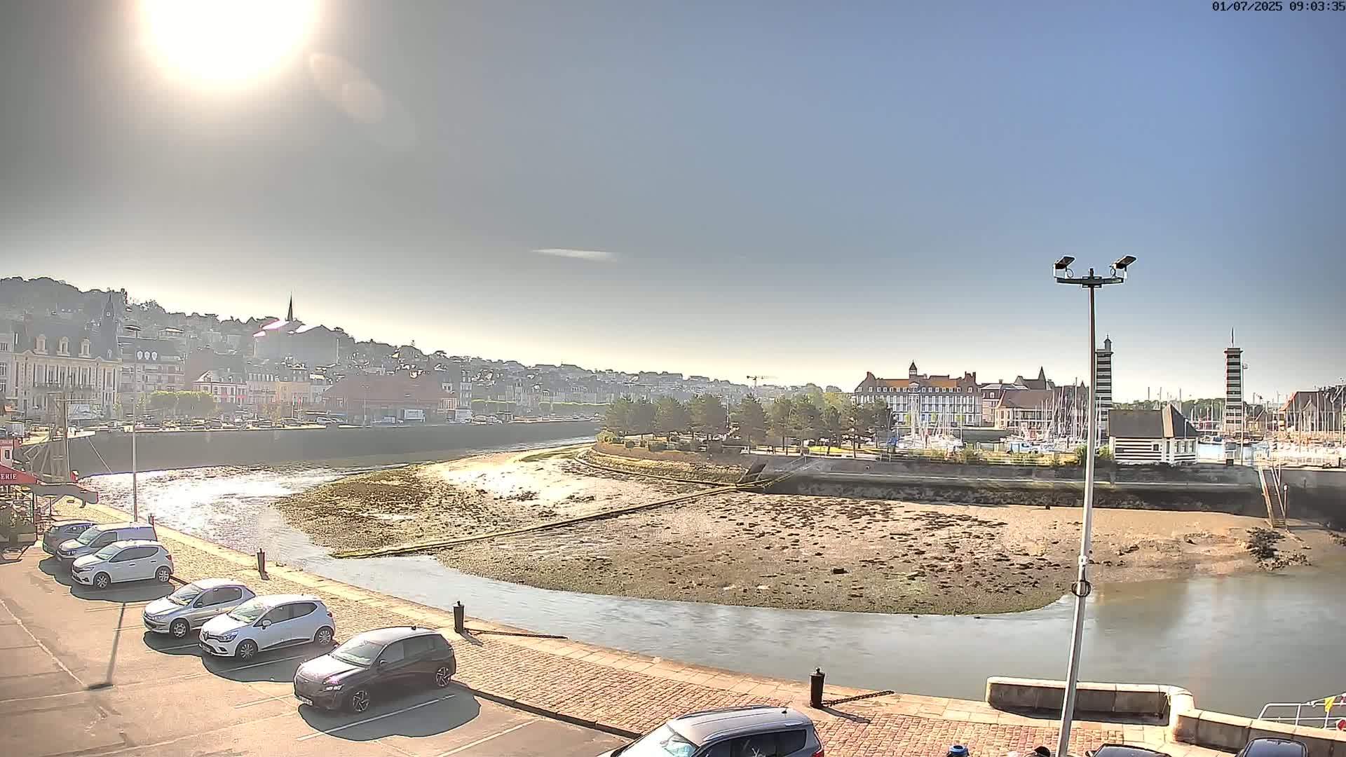 A sunny day reveals a harbor scene with a curving river revealing mudflats at low tide, a parking lot filled with cars, and a town built on a hillside in the background.