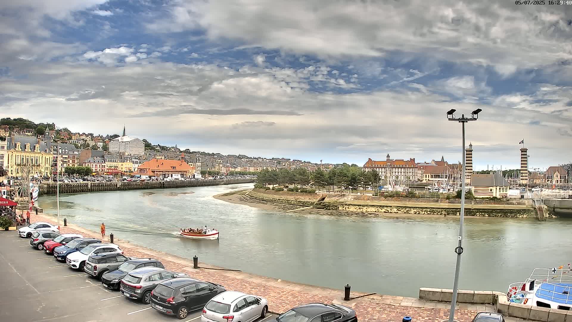 A river flows through a town with buildings lining its banks under a partly cloudy sky, and a small boat is visible on the water.