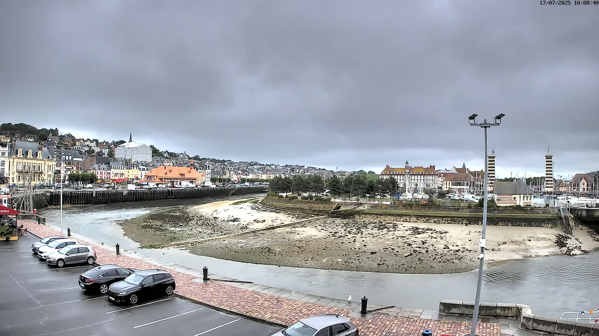 A harbor town under a cloudy sky, with a low tide exposing a wide expanse of muddy seabed, and several cars parked in a lot nearby.