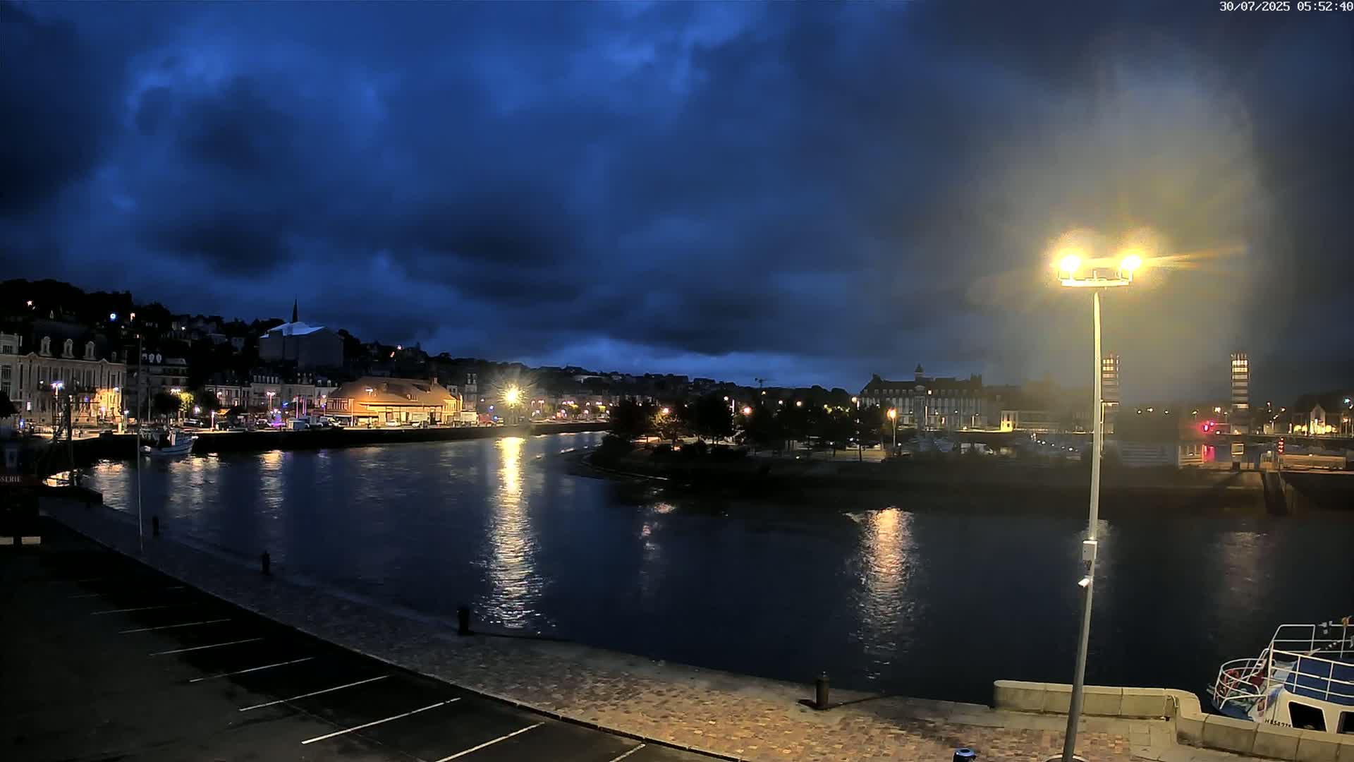 A harbor town under a cloudy sky, with a low tide exposing a wide expanse of muddy seabed, and several cars parked in a lot nearby.