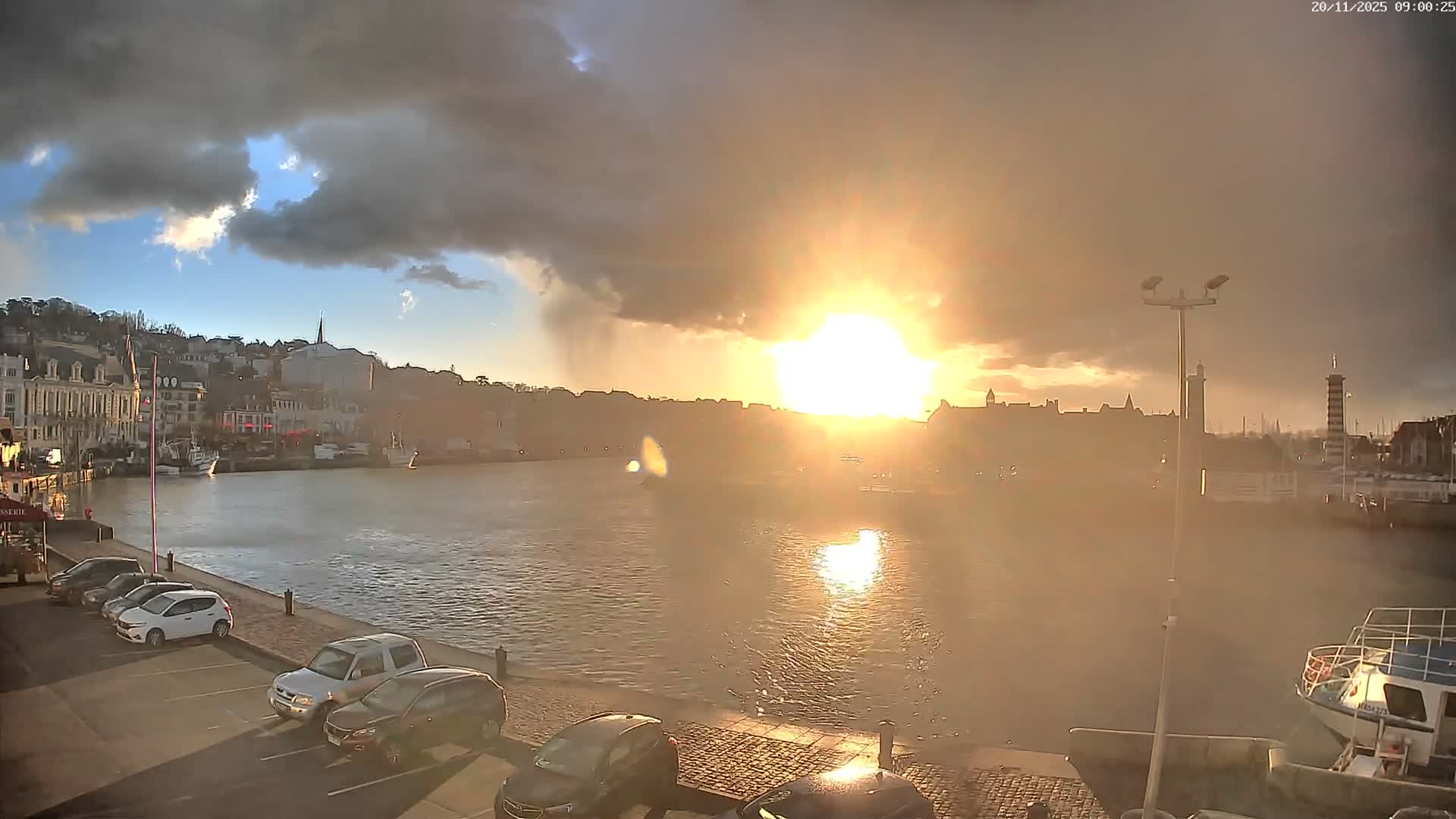 A brilliant sun peeks through dark, dramatic storm clouds, casting a golden light over a bustling harbor or estuary with visible rain falling, illuminating the water, a town's architecture, and parked cars along the waterfront.