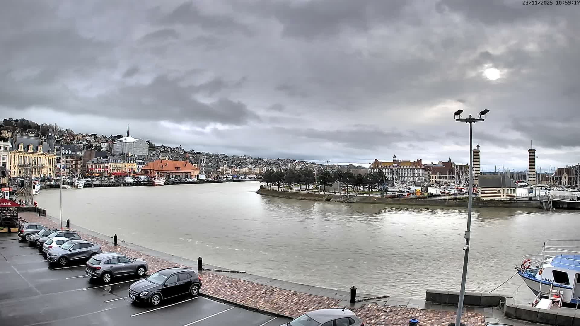 A wide river flows through a densely built coastal town featuring colorful buildings ascending a hillside, a marina with numerous boats and striped lighthouses, all under a heavily overcast sky with the sun barely visible through the clouds.