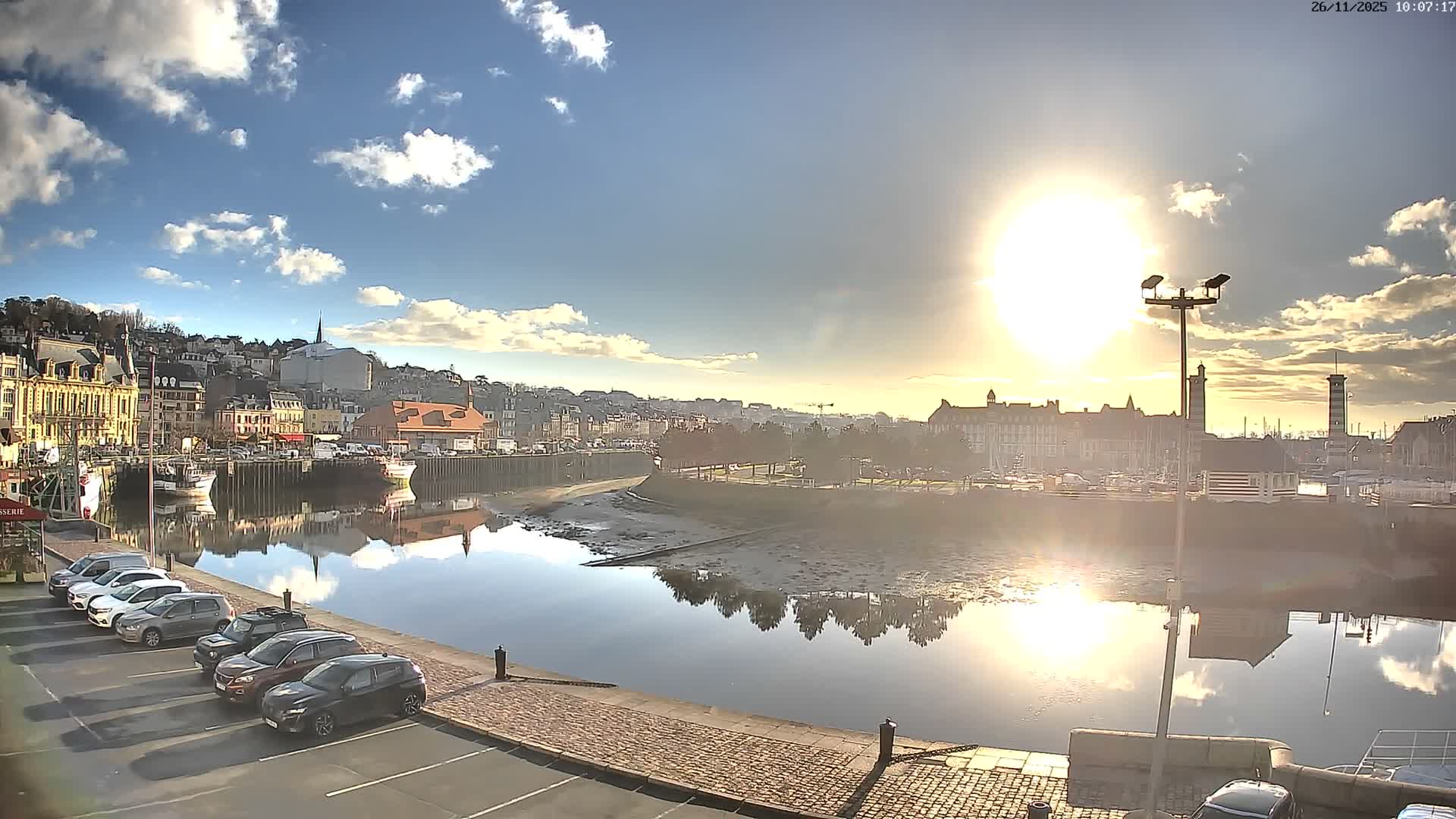 A European coastal town is seen on a sunny day with scattered clouds, featuring a harbor or river with moored boats and parked cars, where the bright sun reflects on the water's surface and reveals muddy banks at low tide.