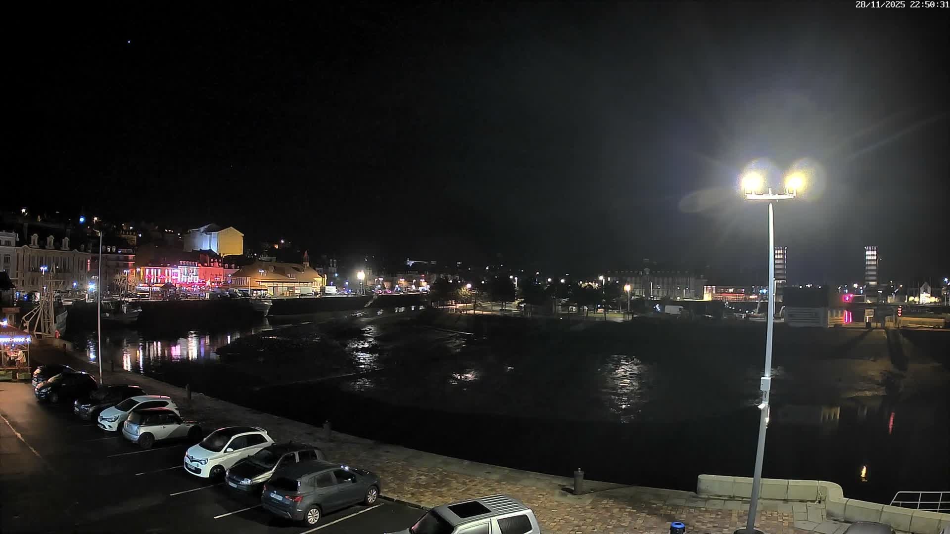 On a clear, dark night, a waterfront town glows with brightly lit buildings and their reflections in the calm water, featuring parked cars along the quay and a prominent streetlamp in the foreground.