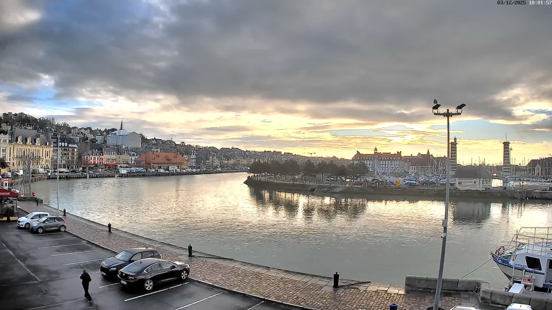 A wide view of a town's waterfront features a winding waterway reflecting the overcast sky's soft golden glow, flanked by buildings, a distant marina with boats, and a wet paved area with parked cars and a person walking.