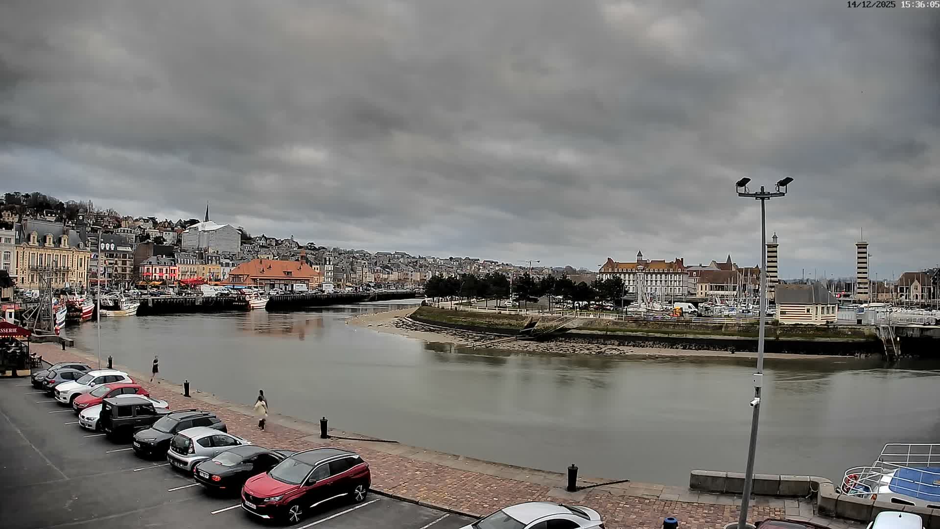A wide view of a town's waterfront features a winding waterway reflecting the overcast sky's soft golden glow, flanked by buildings, a distant marina with boats, and a wet paved area with parked cars and a person walking.