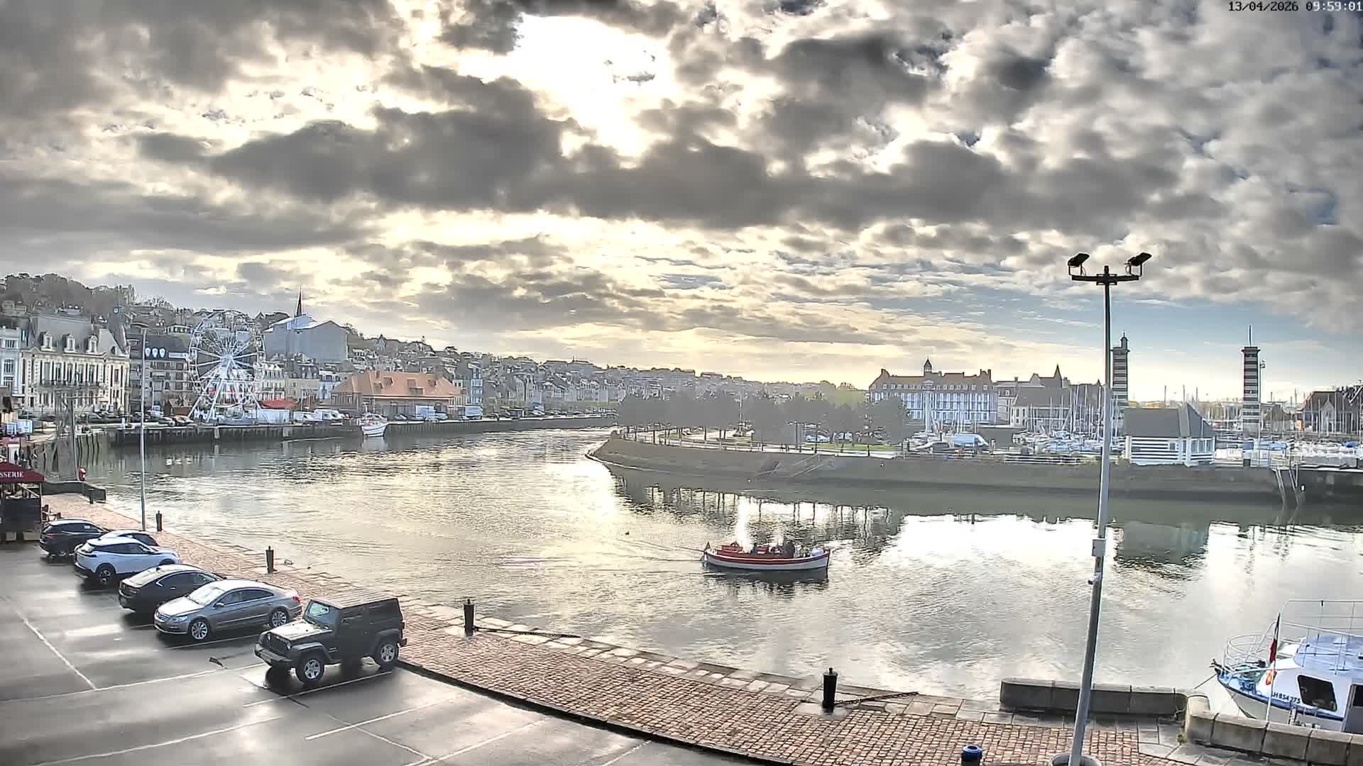 A wide view of a town's waterfront features a winding waterway reflecting the overcast sky's soft golden glow, flanked by buildings, a distant marina with boats, and a wet paved area with parked cars and a person walking.