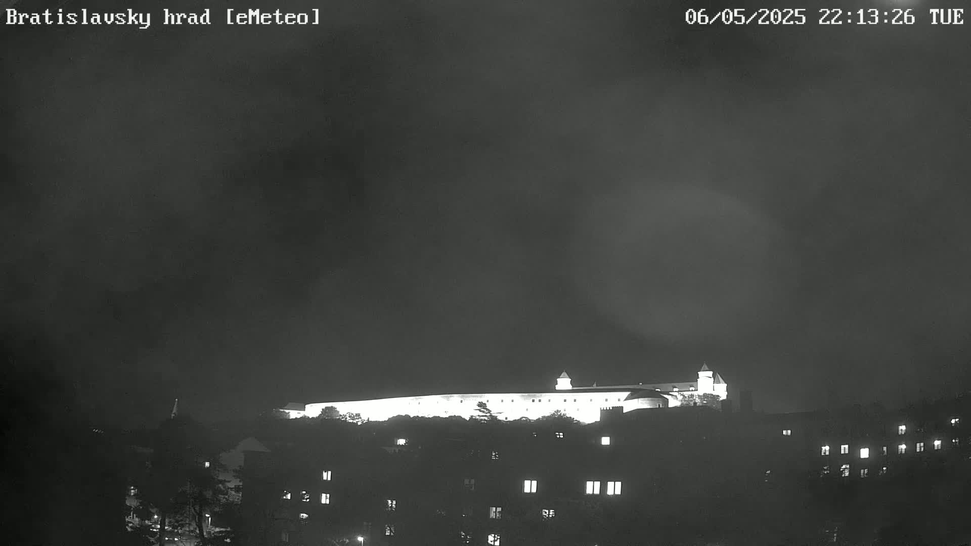 A brightly lit castle sits atop a hill at night under a dark, cloudy sky.