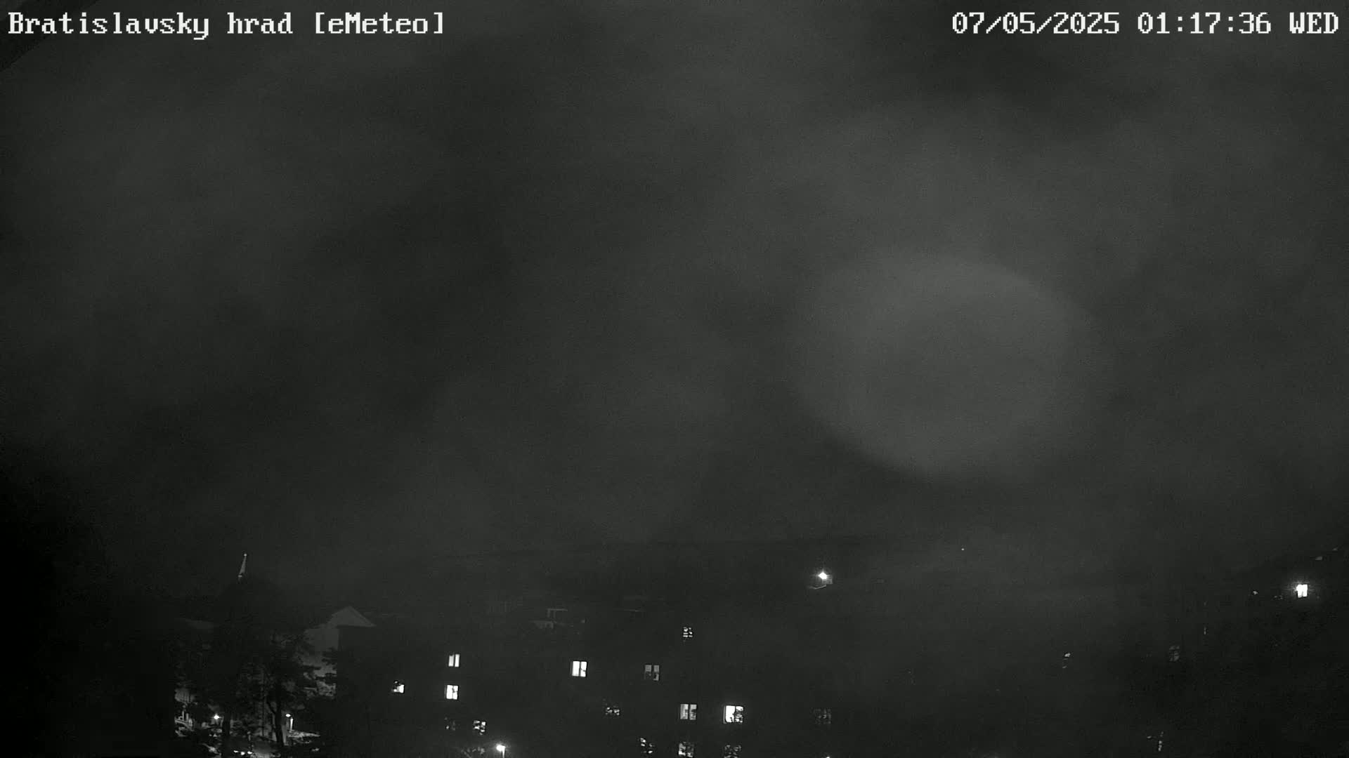A nighttime, hazy view shows dimly lit buildings in the foreground under a dark, overcast sky.