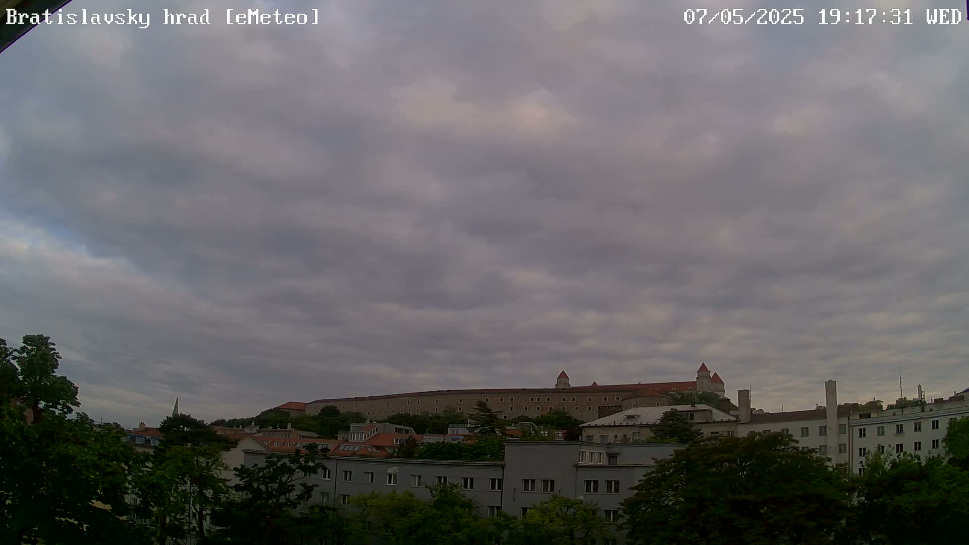 Under a cloudy sky, a large castle sits atop a hill overlooking a city of buildings and green trees.