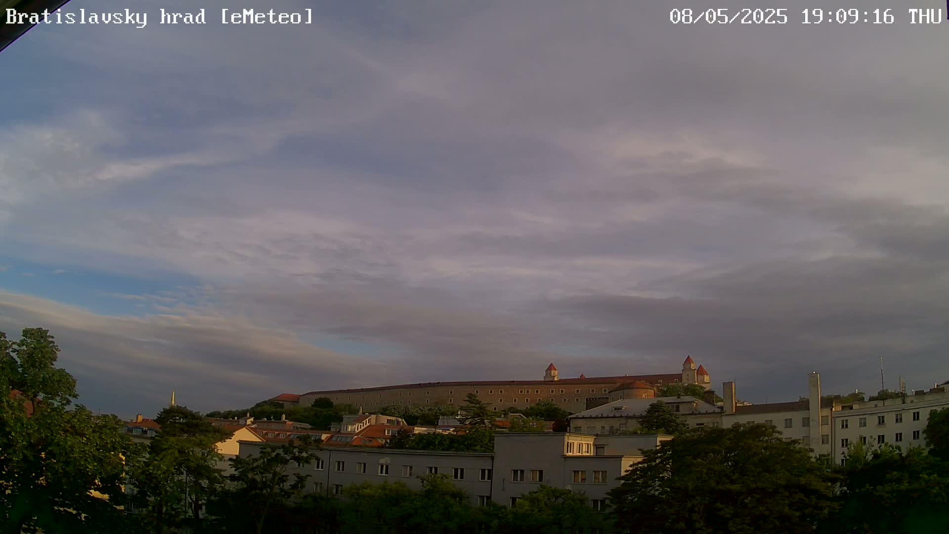 A large castle sits atop a hill behind a city of low-rise buildings under a partly cloudy sky.