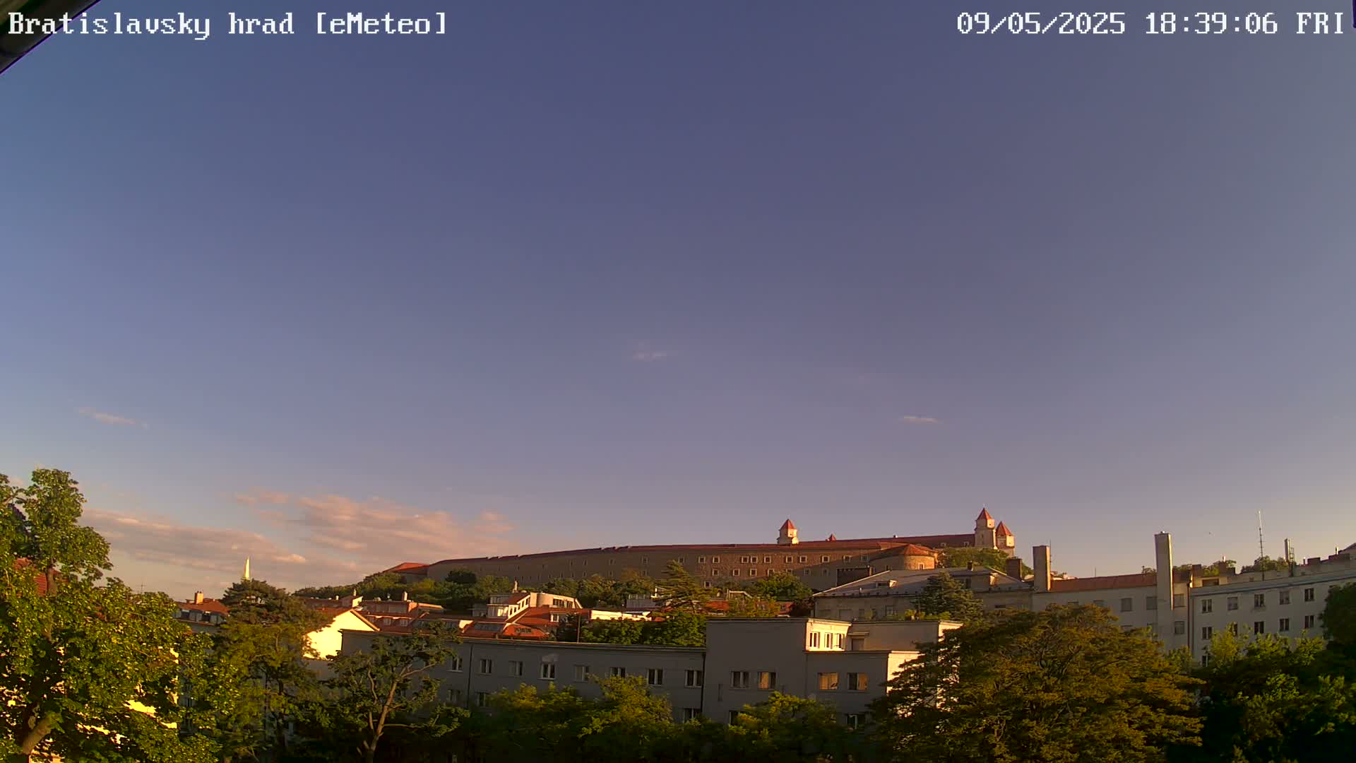 Under a mostly clear, twilight sky, a large castle sits atop a hill overlooking a town of buildings and lush green trees.