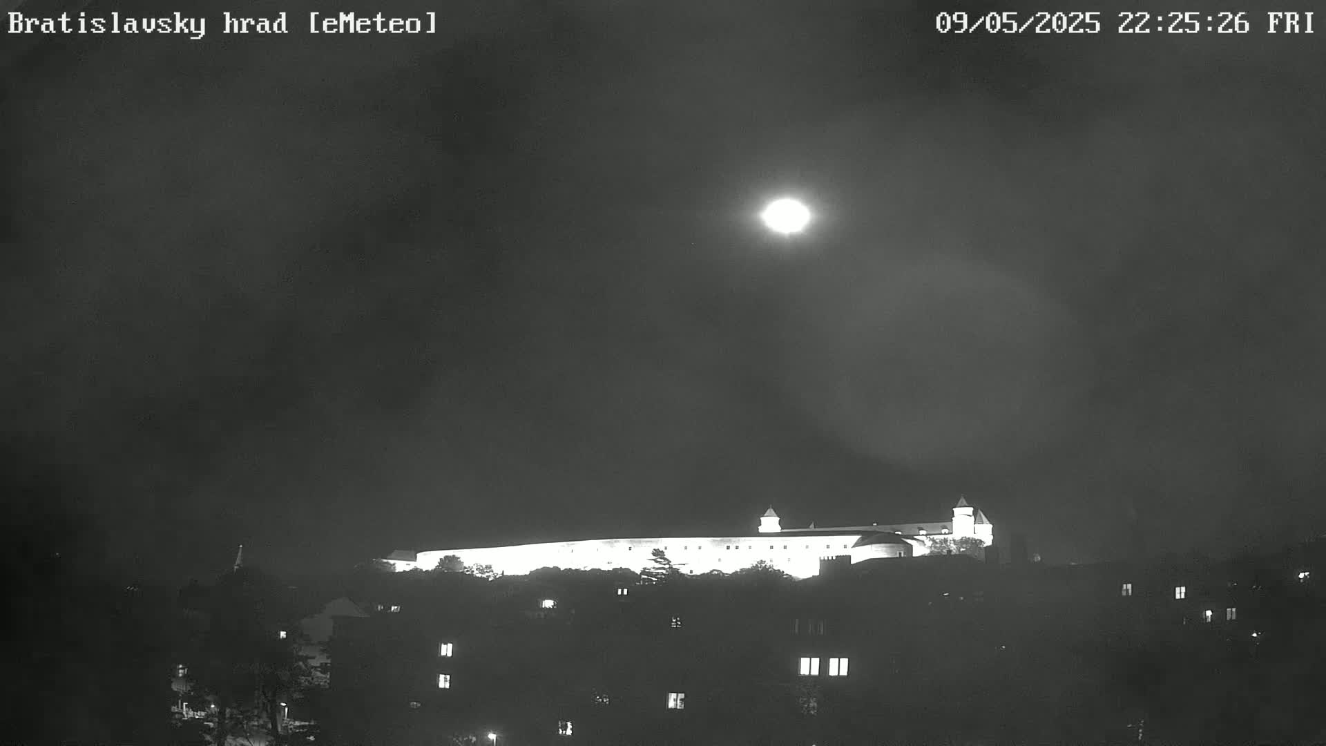 A brightly lit castle sits atop a hill under a night sky with a full moon.