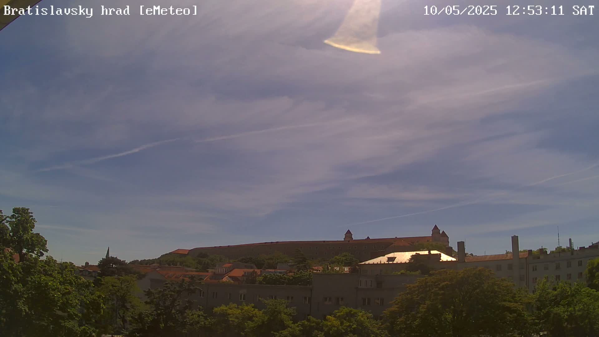 A large castle sits atop a hill under a mostly clear, sunny sky, with green trees in the foreground.