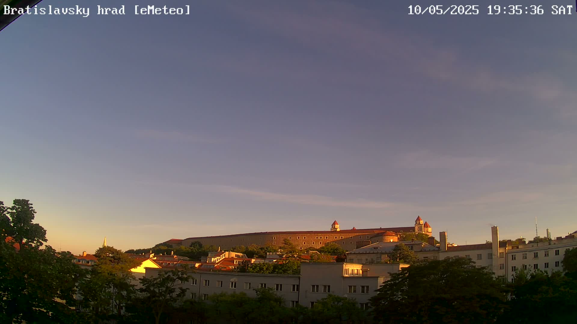 A large stone castle sits atop a hill, overlooking a city of multi-story buildings under a clear, twilight sky.