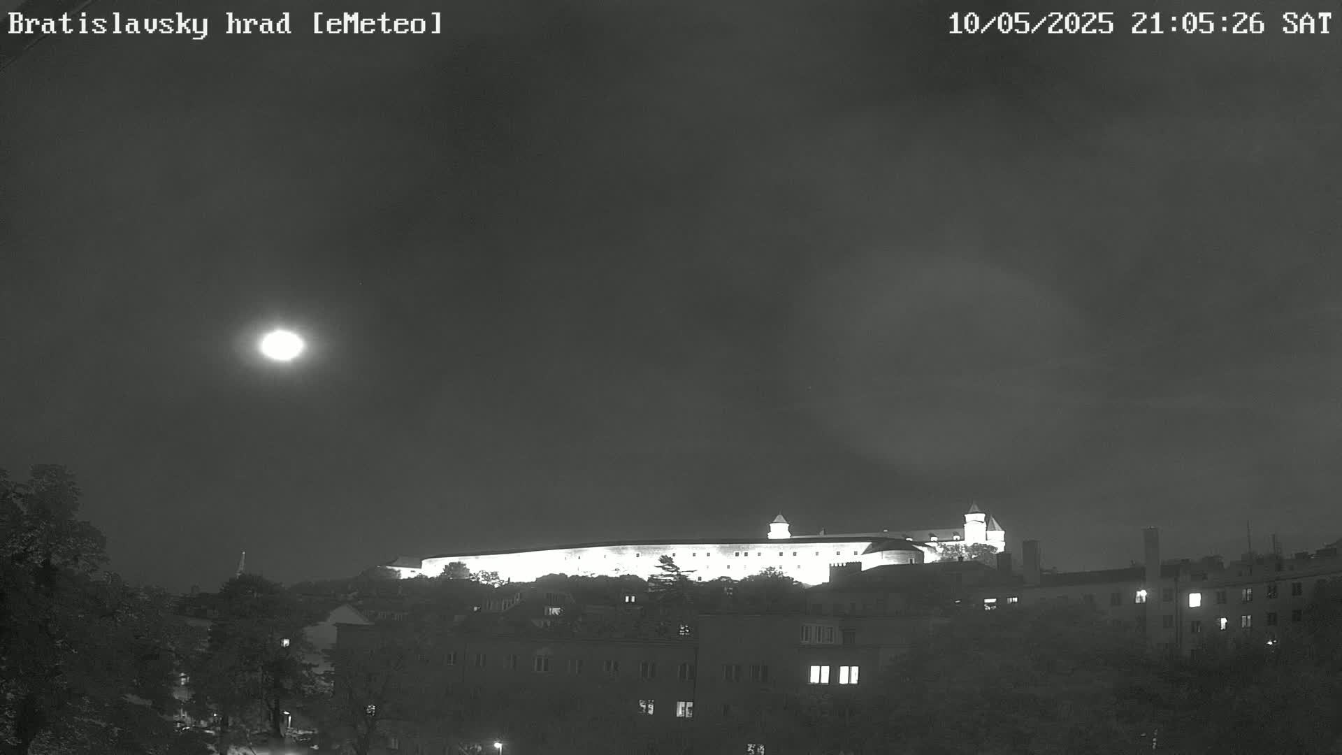 A brightly lit castle sits atop a hill at night under a mostly clear sky and a bright moon.