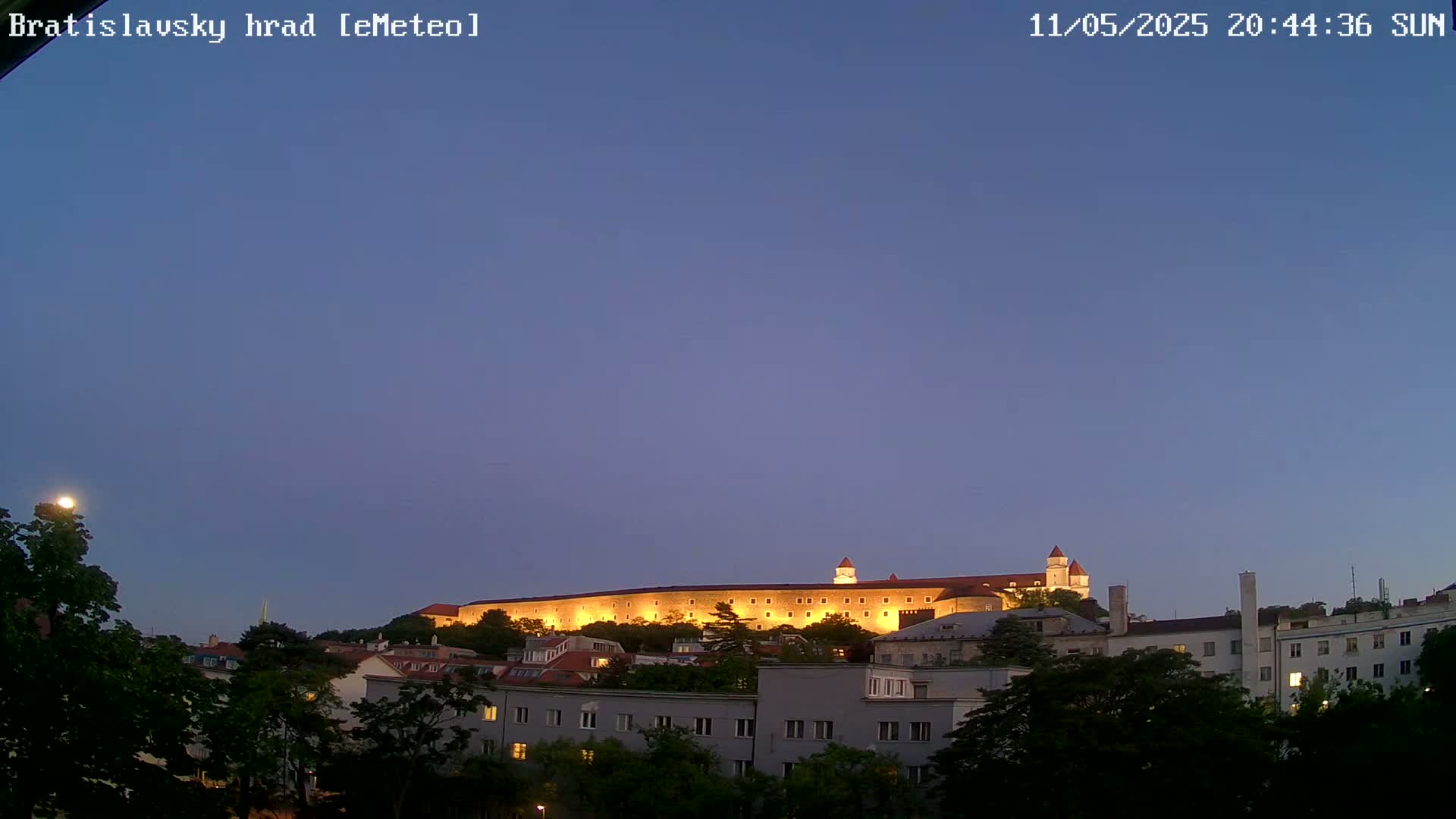 A large illuminated castle sits atop a hill at dusk, overlooking a city with buildings and trees in the foreground under a clear twilight sky.