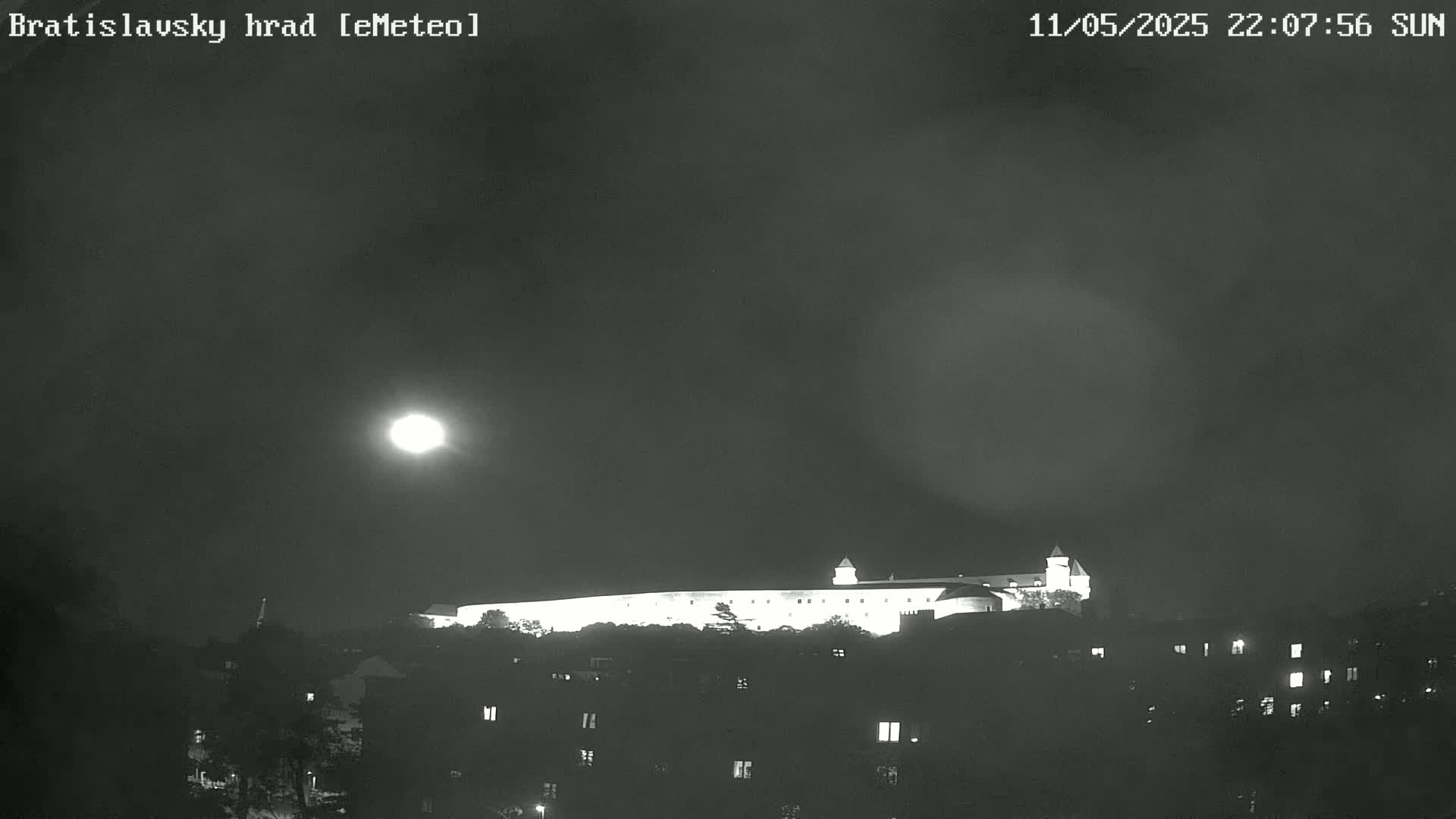 A brightly lit castle sits atop a hill under a mostly cloudy night sky illuminated by a bright moon.