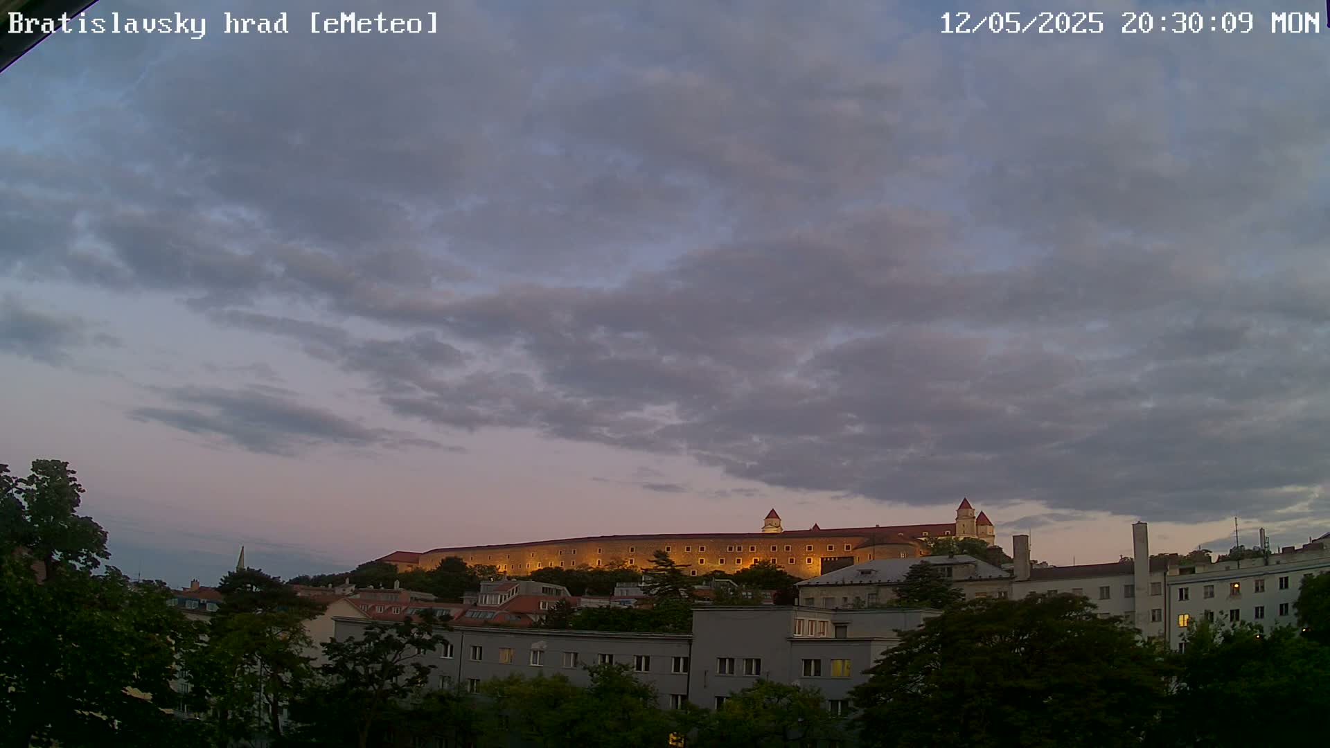 A large castle is illuminated at dusk under a cloudy sky, situated behind a row of buildings and trees.