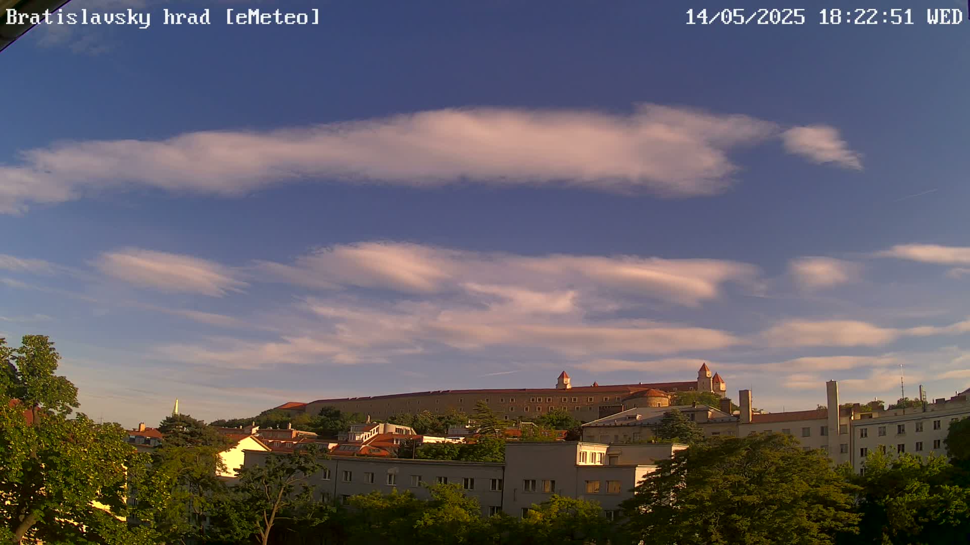 A long, low, red-roofed castle sits atop a hill behind a town of low buildings under a mostly clear sky with wispy clouds.