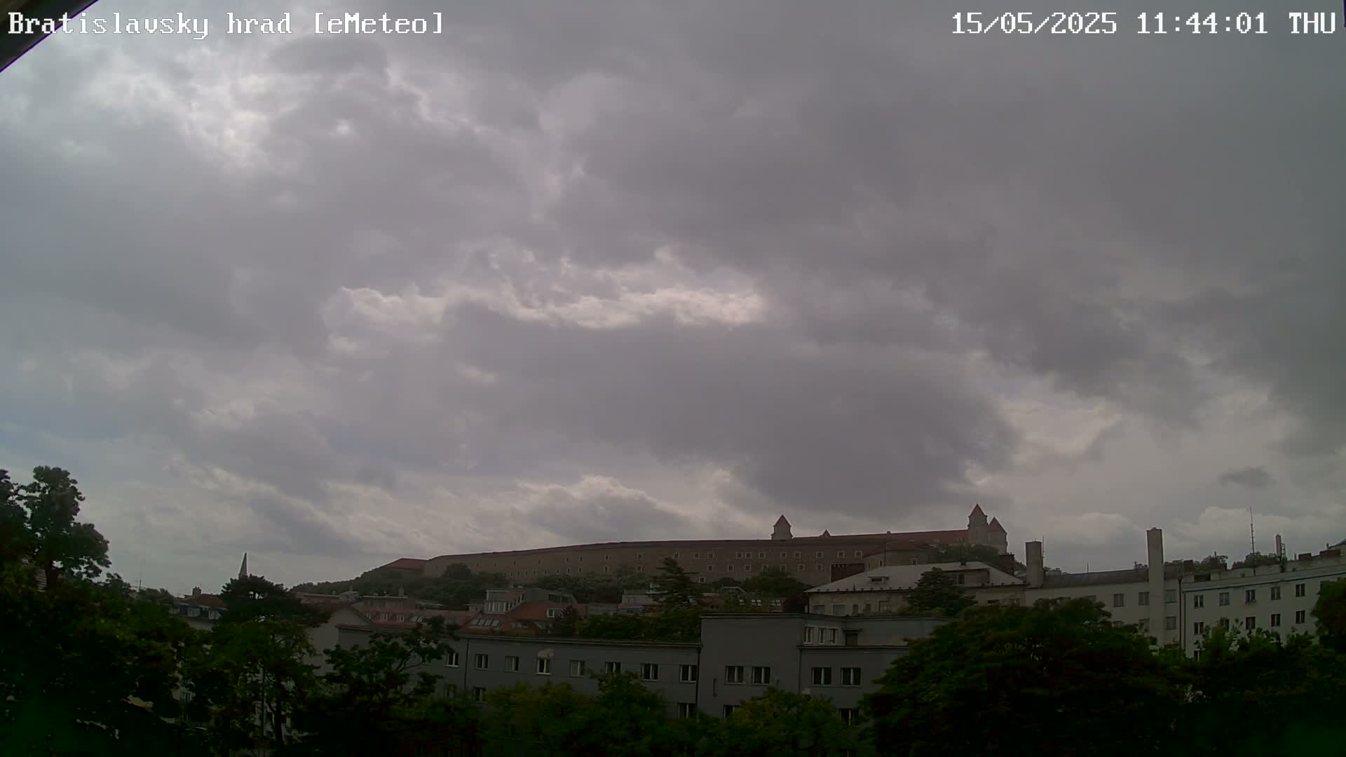 A large stone castle sits atop a hill behind a row of buildings under a cloudy sky.