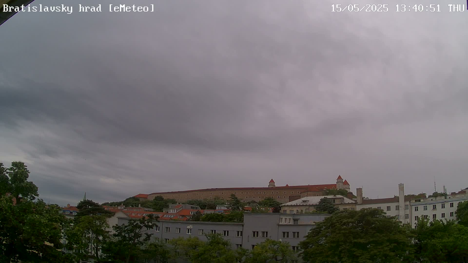 A large stone castle sits atop a hill behind a row of buildings under a cloudy sky.