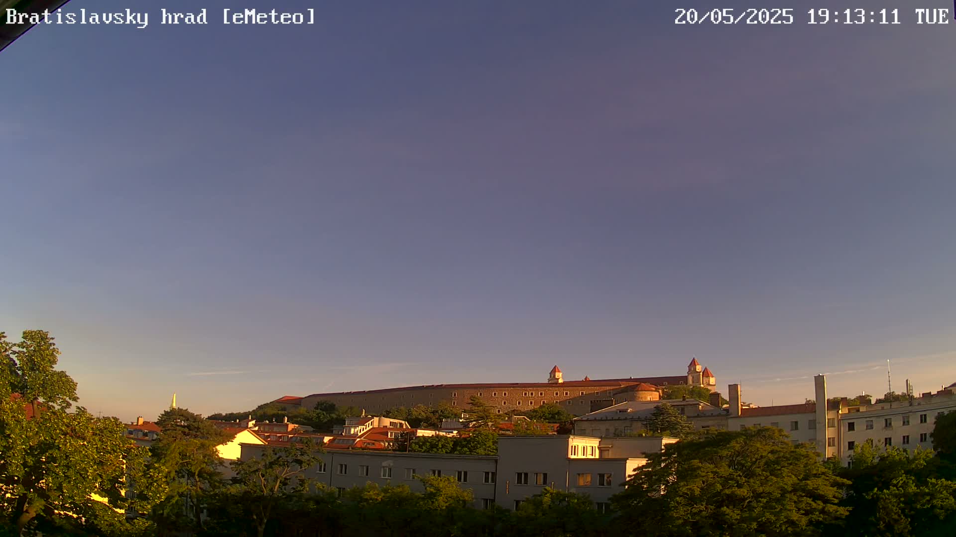 A long, stone castle sits atop a hill overlooking a city of low-rise buildings under a clear, twilight sky.