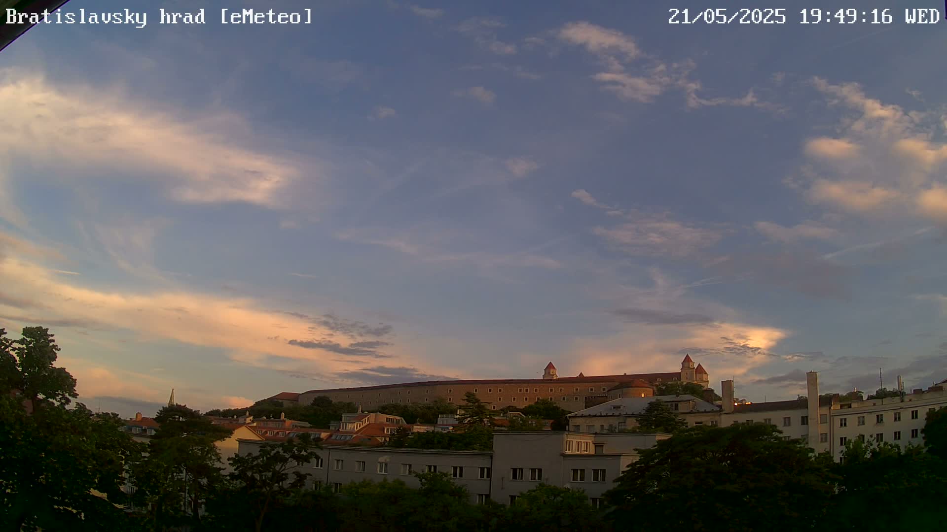 A large castle sits atop a hill, partially obscured by trees and buildings in the foreground, under a partly cloudy, sunset sky.
