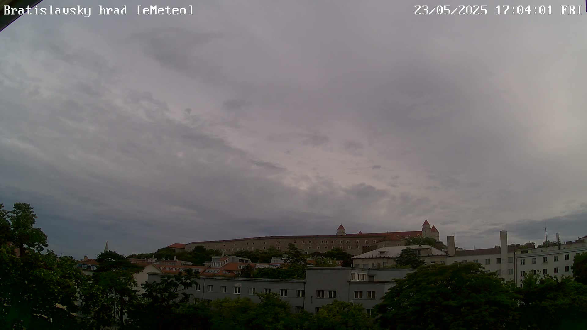 A long, stone castle sits atop a hill in the distance, partially obscured by an overcast sky, behind a row of buildings and green trees.