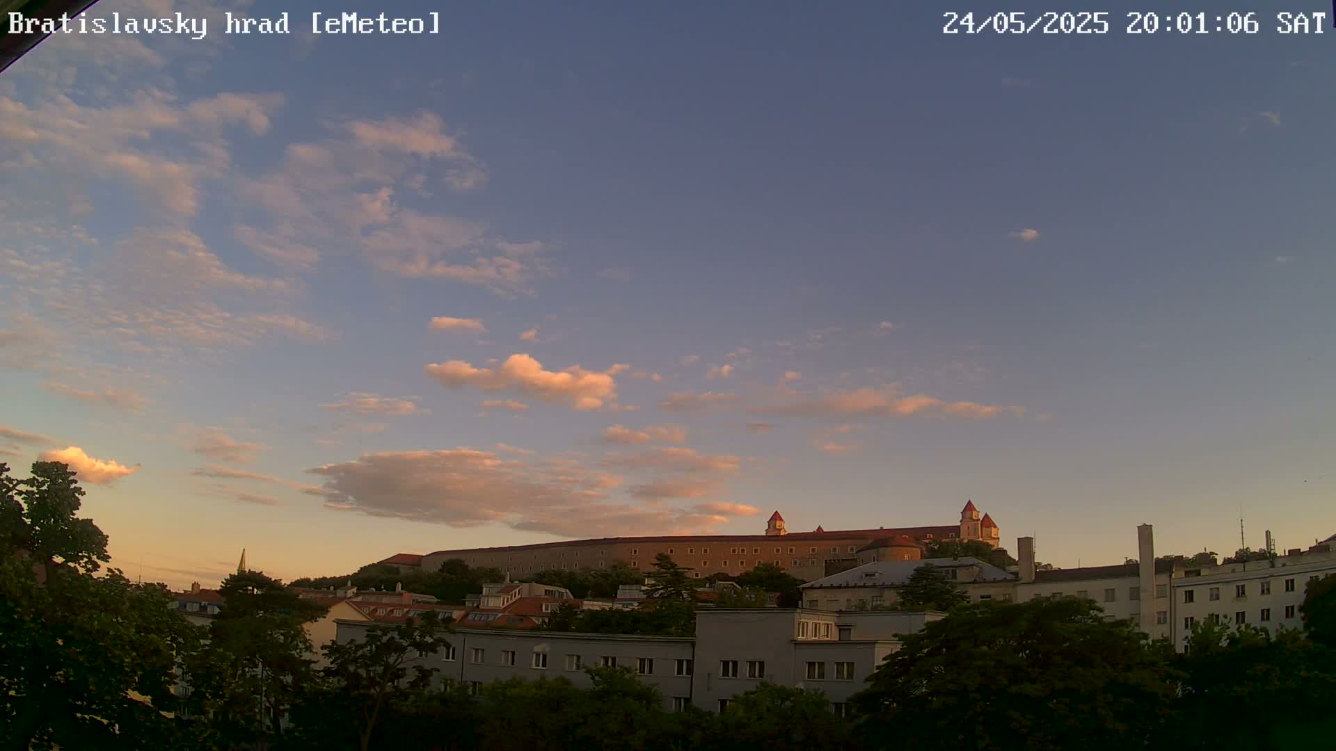 A large castle sits atop a hill under a partly cloudy, pinkish sunset sky, with buildings and trees in the foreground.