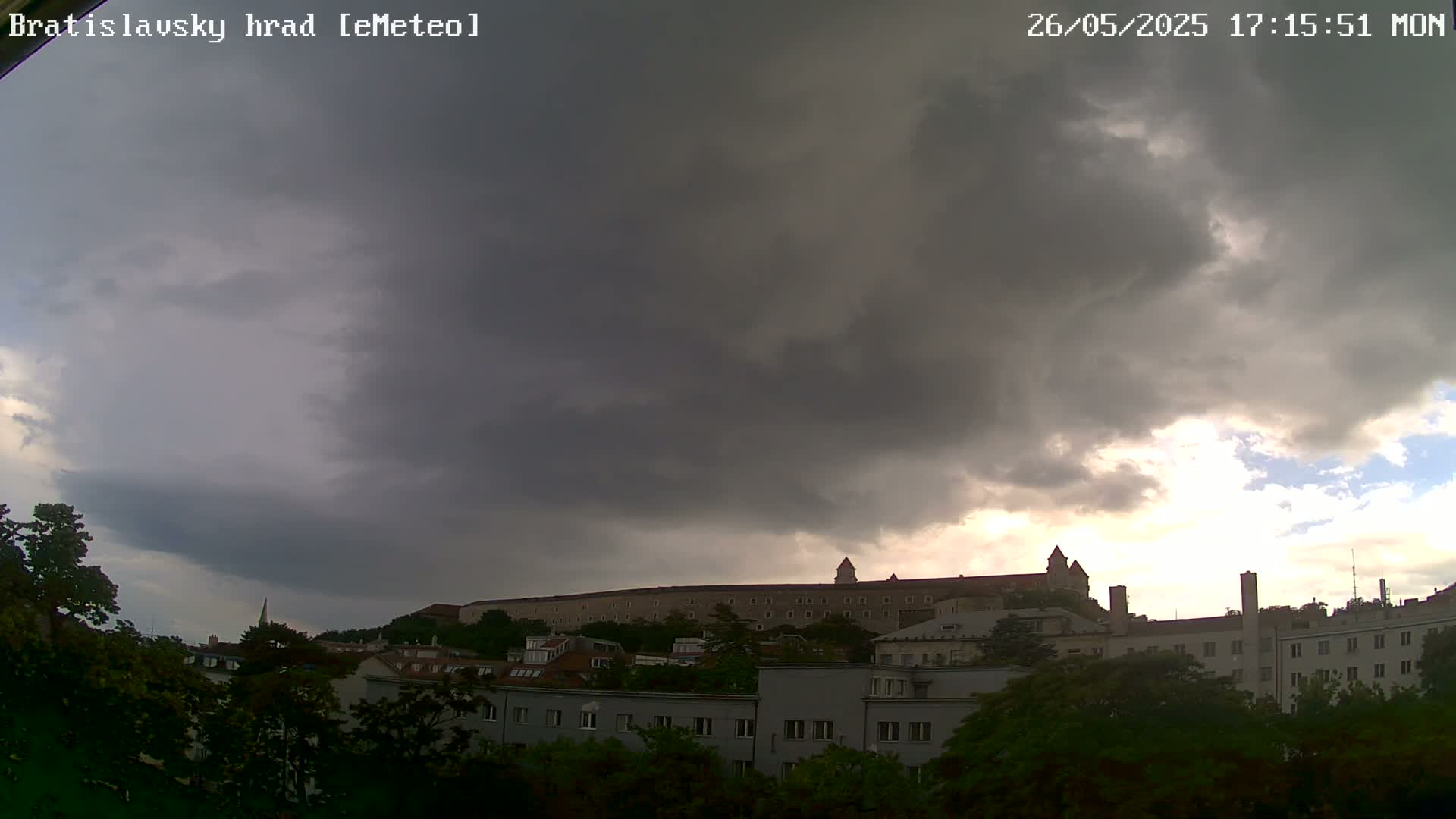 A large, imposing castle sits atop a hill, partially obscured by dark, stormy clouds under which a city of buildings and green trees stretches out.