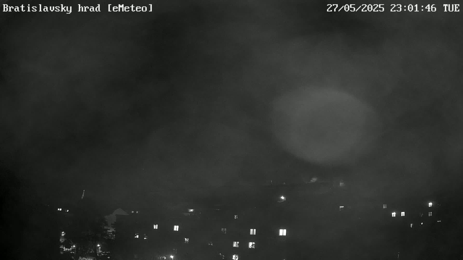 A nighttime, foggy view shows dimly lit buildings in the foreground and a hazy sky above.