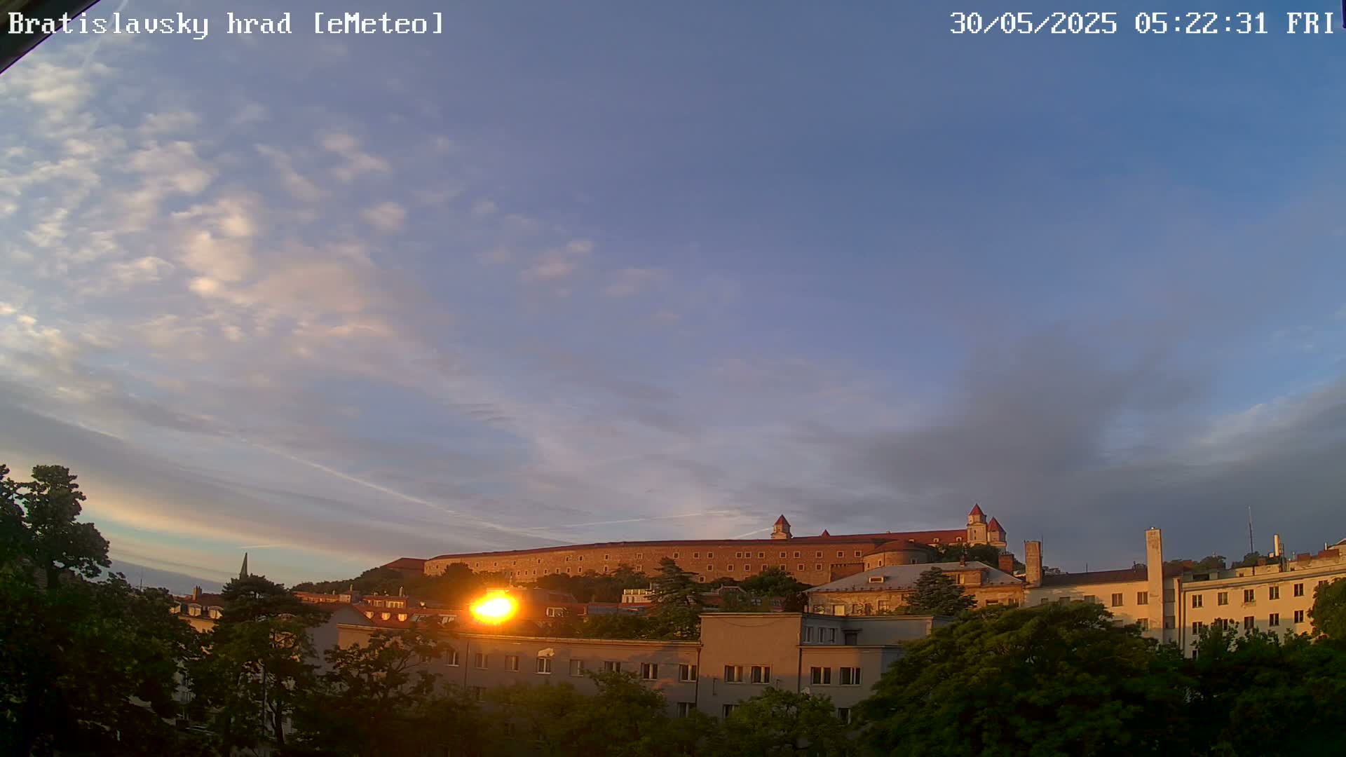 A long, sprawling castle sits atop a hill, visible through a partly cloudy sunrise sky above a cityscape of low buildings and lush green trees.