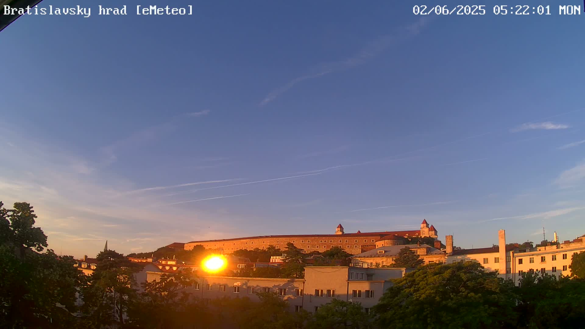 A large castle sits atop a hill overlooking a city at sunrise under a clear, pale blue sky.