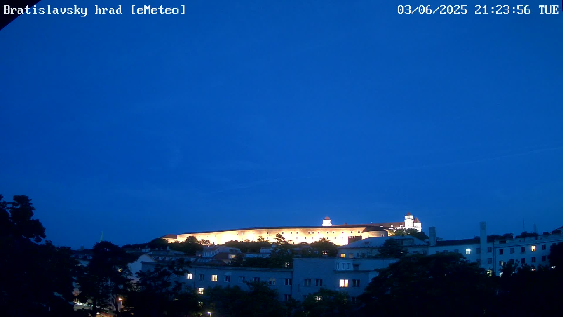 A brightly lit castle sits atop a hill at night, under a clear, dark blue sky, overlooking a town with buildings and trees.