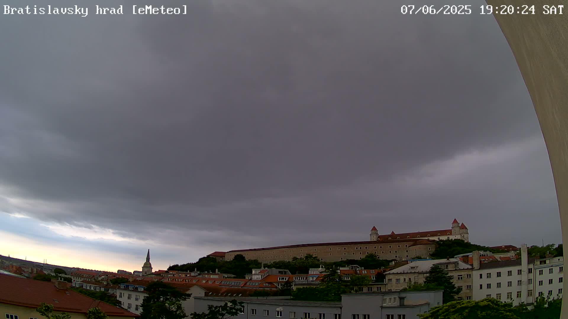 A large, imposing castle sits atop a hill overlooking a city under a mostly cloudy, overcast sky.